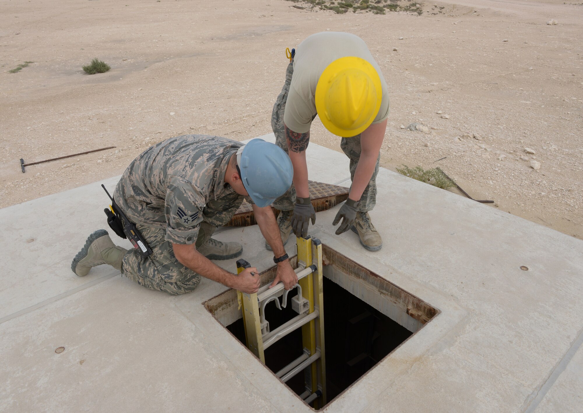 Senior Airman Gary Boston and Airman 1st Class Tony Martin lower a ladder into a manhole at Al Udeid Air Base, Qatar, Mar. 10, 2014. Inside the manhole Airmen from the 379th Expeditionary Communications Squadron cable and antenna systems shop, or Cable Dawgs, maintain fiber optic cable used to run communication lines throughout the entire base. Both Airmen are 379th ECS cable and antenna systems technicians.  Boston is deployed from Barksdale Air Force Base, La., and an Oscoda, Mich., native. Martin is deployed from Eglin AFB, Fla., and a Tucson, Ariz., native. (U.S. Air Force photo/Senior Airman Hannah Landeros) 