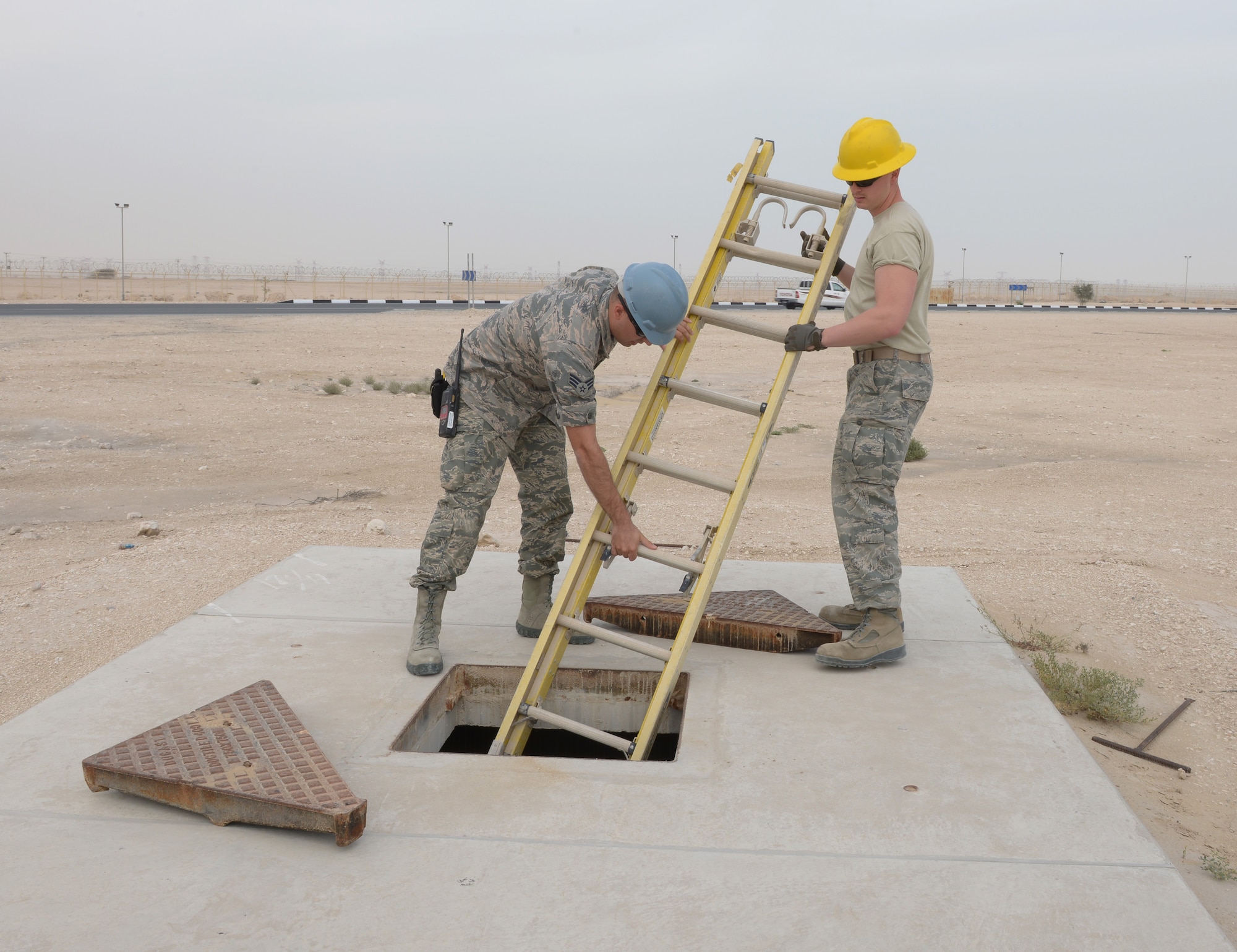 Senior Airman Gary Boston and Airman 1st Class Tony Martin lower a ladder into a manhole at Al Udeid Air Base, Qatar, Mar. 10, 2014. Inside the manhole Airmen from the 379th Expeditionary Communications Squadron cable and antenna systems shop, or Cable Dawgs, maintain fiber optic cable used to run communication lines throughout the entire base. Both Airmen are 379th ECS cable and antenna systems technicians.  Boston is deployed from Barksdale Air Force Base, La., and an Oscoda, Mich., native. Martin is deployed from Eglin AFB, Fla., and a Tucson, Ariz., native. (U.S. Air Force photo/Senior Airman Hannah Landeros) 