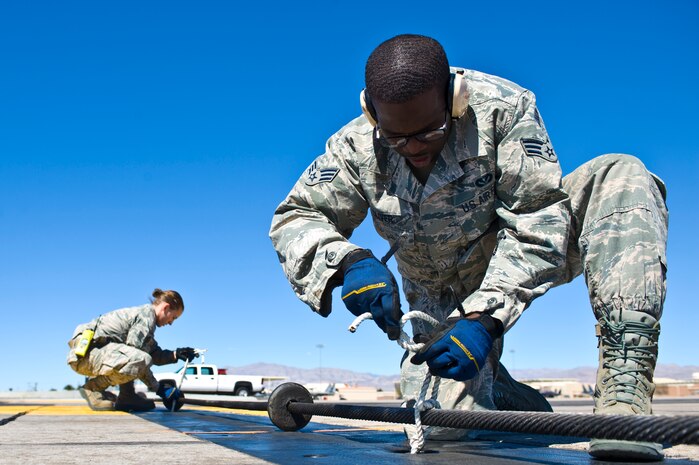U.S. Air Force Senior Airman Demarcus Oliver, 99th Civil Engineer Squadron firefighter, inspects and ties rope around a BAK-12 aircraft arresting barrier on the flightline March 12, 2014, at Nellis Air Force Base, Nev. Rope is used to keep the barrier in place for aircraft landings. (U.S. Air Force photo by Senior Airman Christopher Tam)