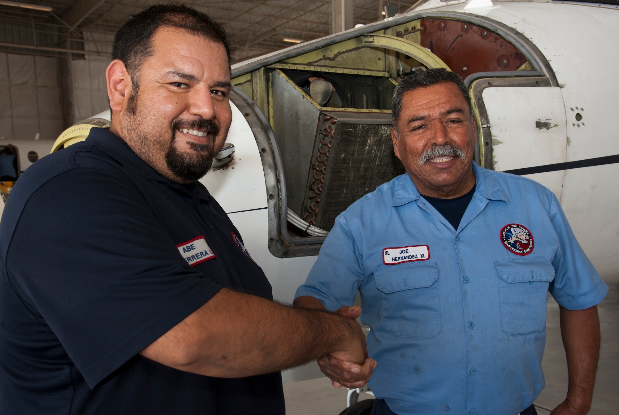 Abe Herrera, left, and Joe Hernandez, right, 47th Maintenance Directorate aircraft maintainers, shake hands and pose for a photo in the T-1 Hangar at Laughlin Air Force Base, Texas, March 21, 2014. Herrera performed the Heimlich maneuver on Hernandez, saving him from choking. (U.S. Air Force photo/Senior Airman Nathan Maysonet)