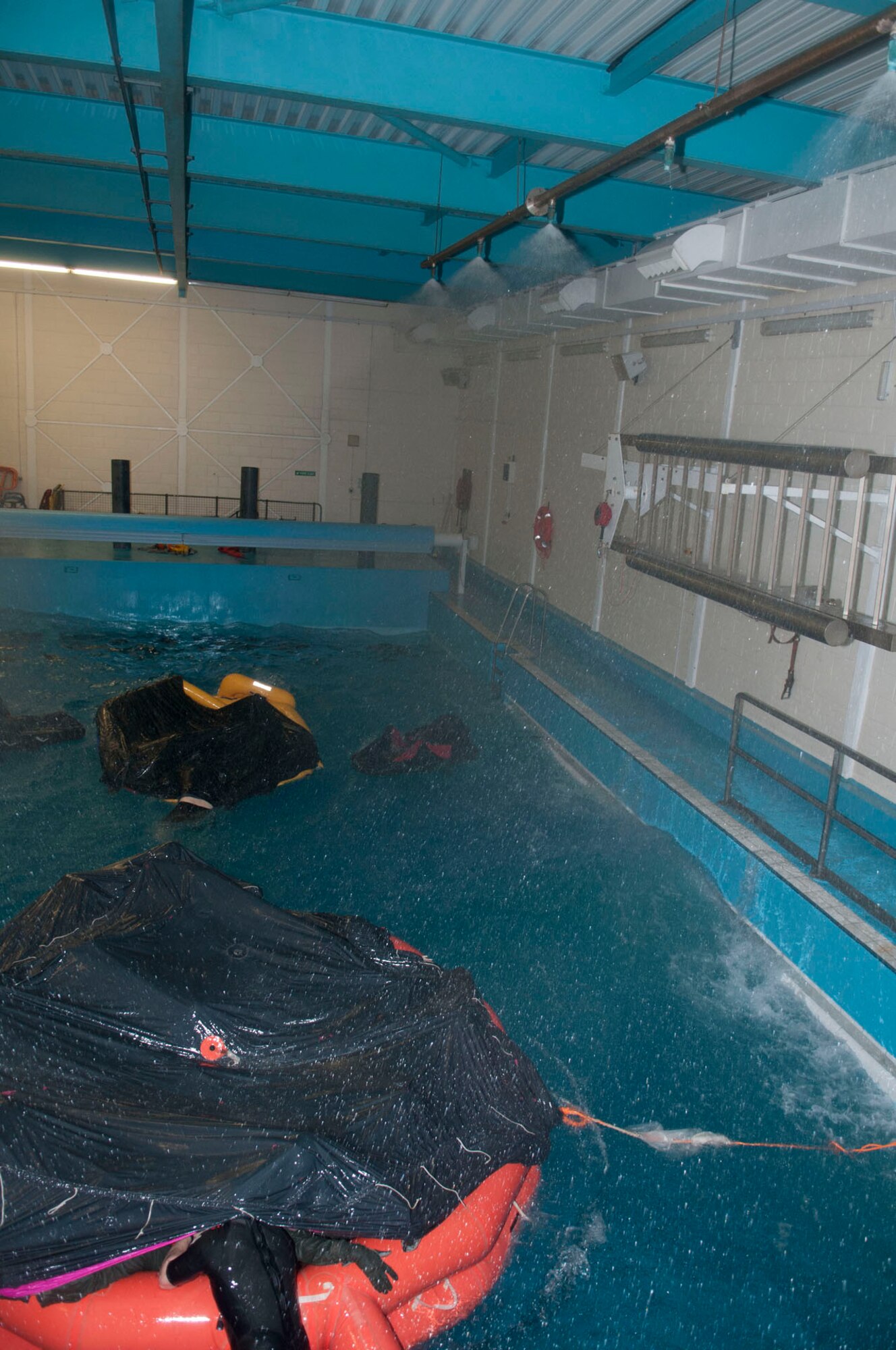 Sprinklers rain down on top of aircrew from Team Mildenhall and RAF Lakenheath's Liberty Wing as they move a life raft to evasion mode during water survival training March 20, 2014, at Lowestoft College in Norfolk, England. The special-purpose pool can recreate extreme weather conditions, including 30-knot winds and 6-foot waves, for aircrew to learn from Survival, Evasion, Resistance and Escape specialists. (U.S. Air Force photo by Karen Abeyasekere/Released)