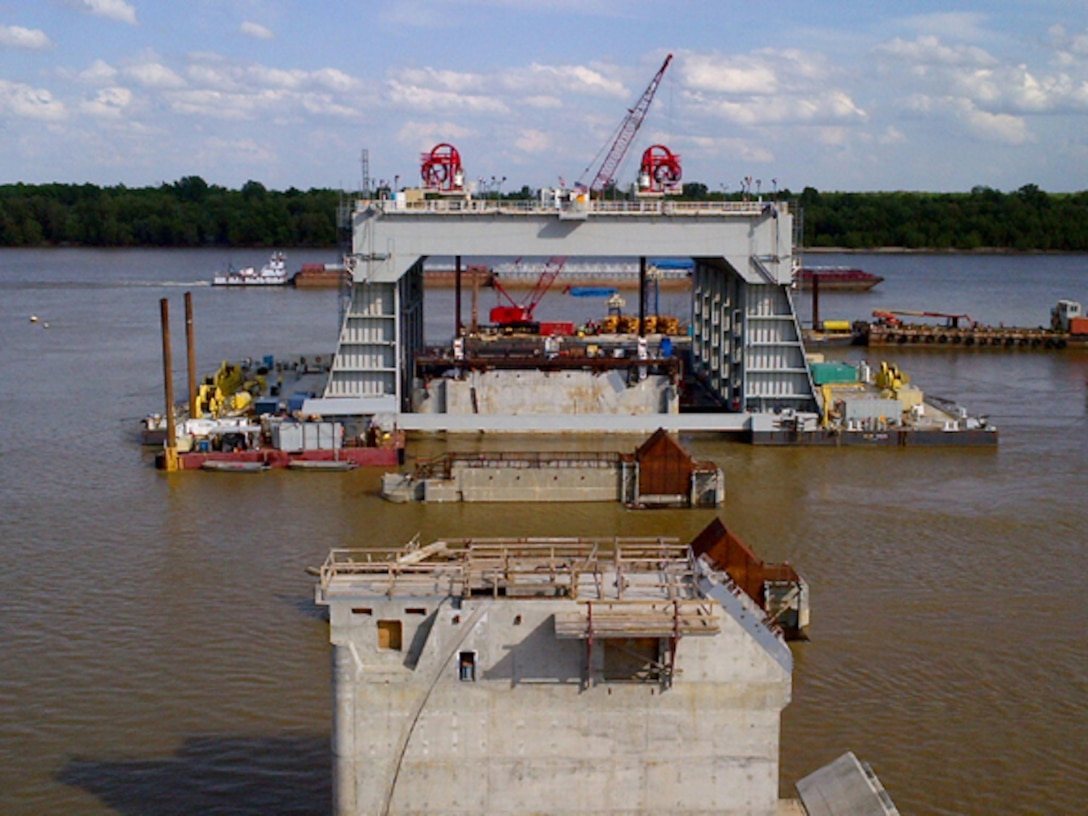 The catamaran barge (background) is used to set shells that comprise the dam. 

The catamaran barge lifts and carries the large precast concrete shells with attached lifting frames on the Ohio River to the shell’s final set-down location.