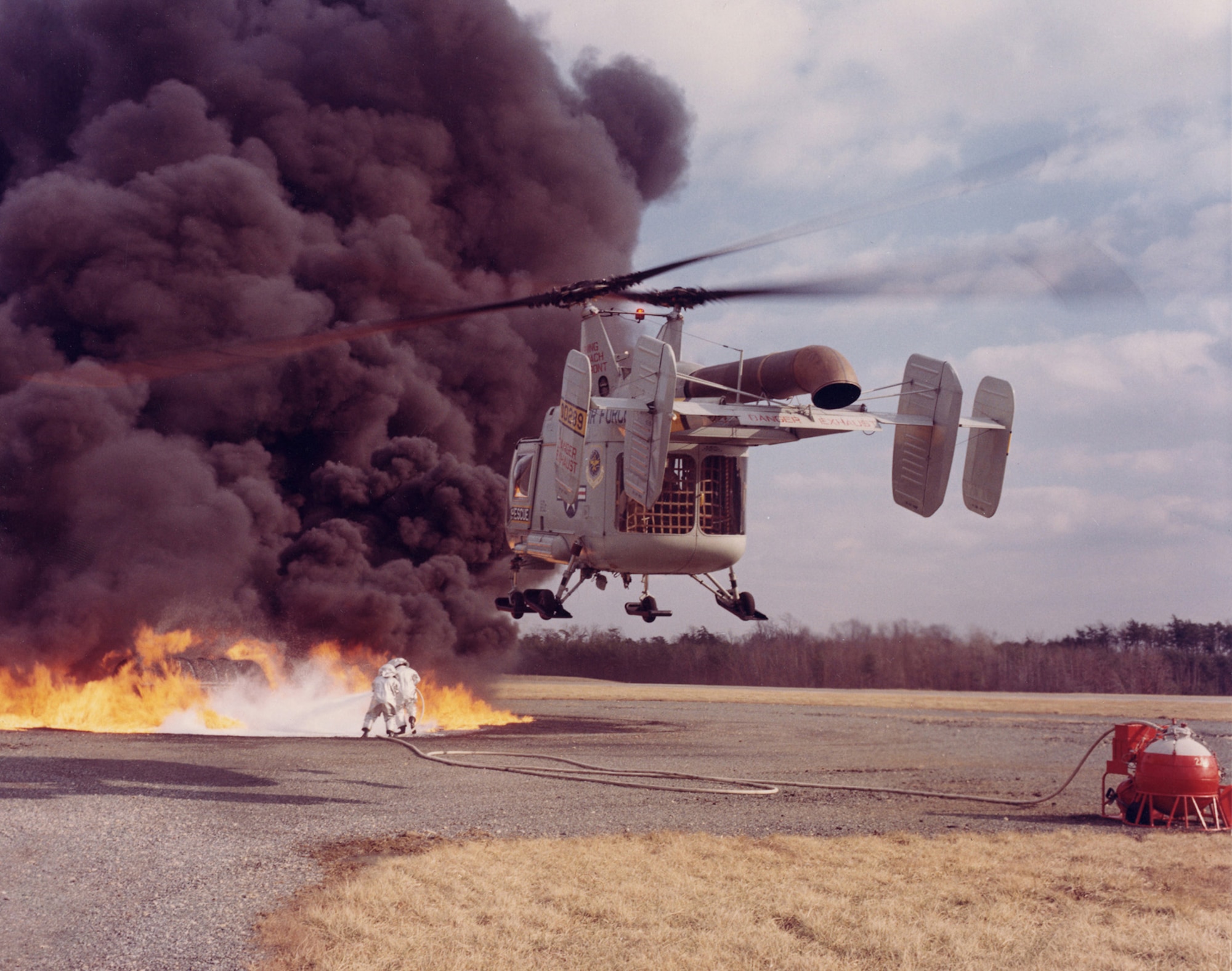 HH-43 crew practicing Local Base Rescue/Firefighting. Downwash air from the rotors opened a path for rescuers to spray foam from the red and white fire suppression kit in the lower right. (U.S. Air Force photo)