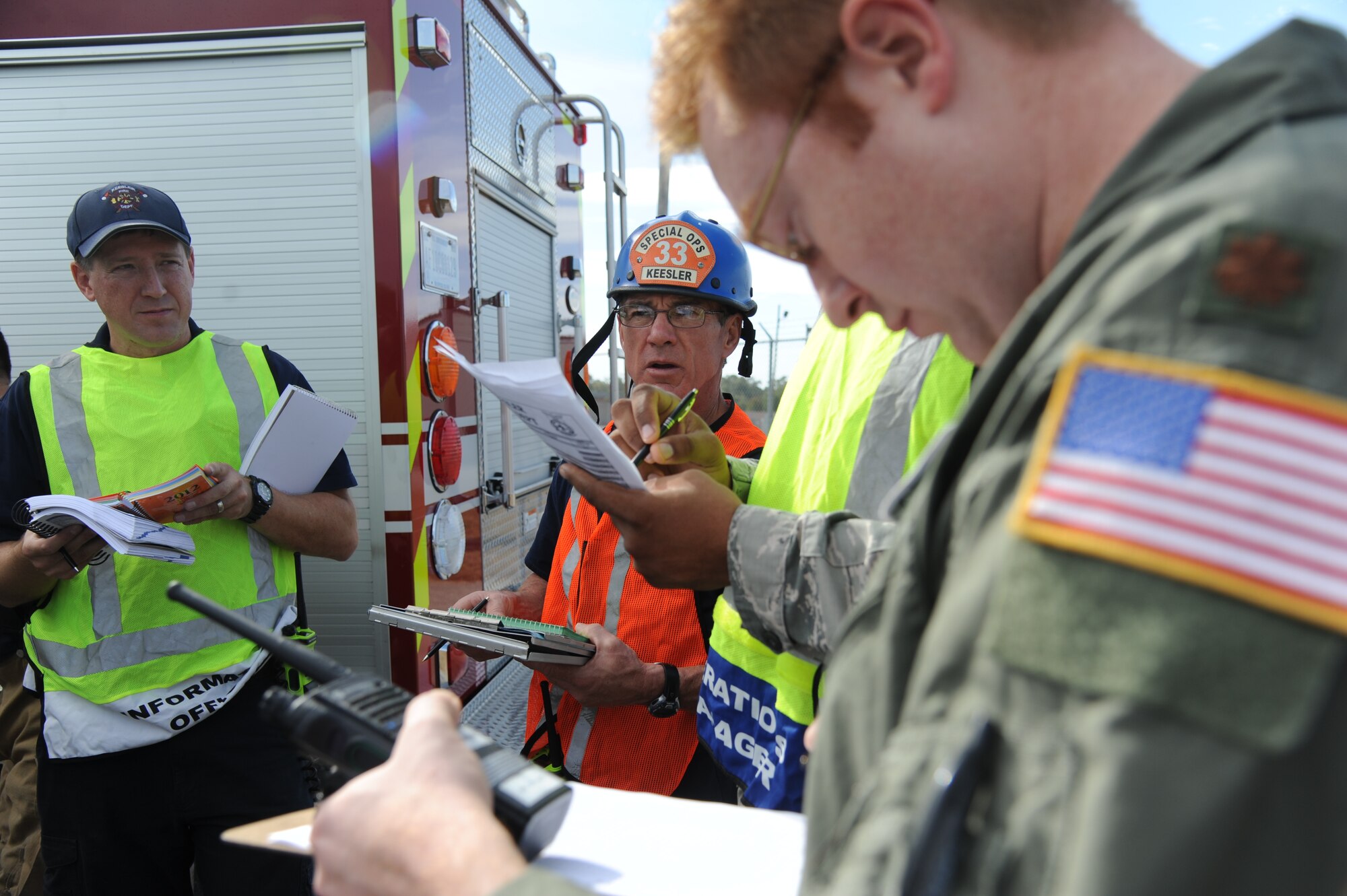 Rusty Bell, Keesler firefighter special operations, (center), discuss procedures for patient treatment and transport with Chris Oliver, Keesler firefighter information officer, and Maj. Shane Steiner, 81st Aerospace Medicine Squadron field response team chief, during a hazardous material exercise scenario Nov. 5, 2014, at the Taylor Logistics Center, Keesler Air Force Base, Miss.  The exercise scenario began with an Airman accidentally hitting a 55-gallon drum of sulfuric acid with a forklift and knocking several more drums off of a truck causing the liquid to splash onto other Airmen.  Immediately, the Airmen simulated skin and eye burns from exposure to the agent, while additional Airmen on the loading dock started coughing and experiencing shortness of breath. (U.S. Air Force photo by Kemberly Groue)