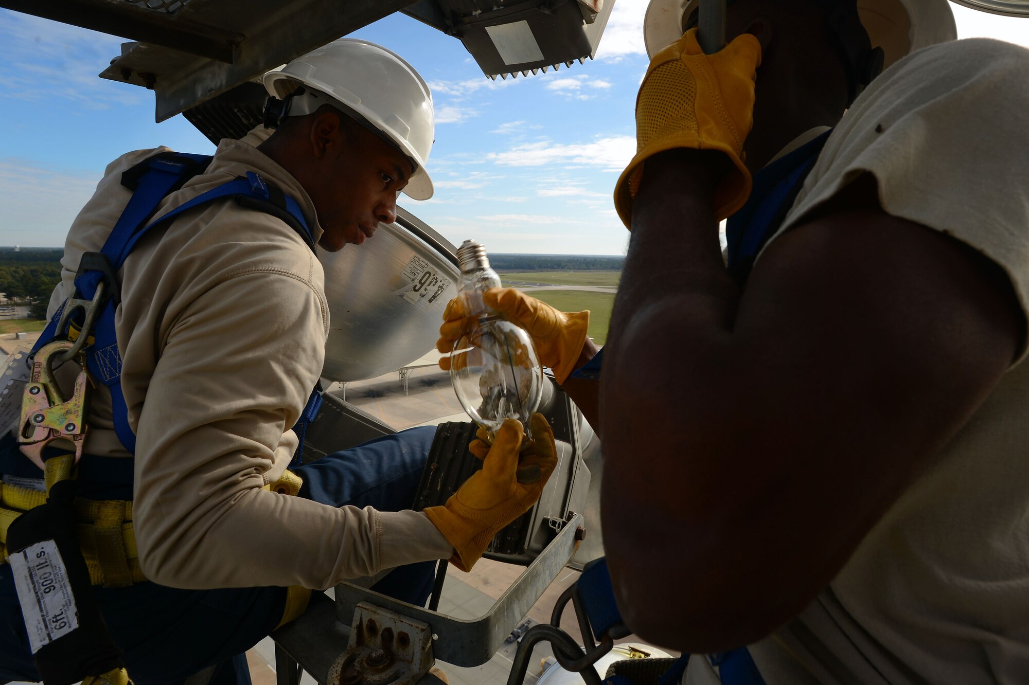 U.S. Air Force Airman 1st Class Ernest Kuma, 20th Civil Engineer Squadron electrical systems apprentice, hands a light bulb to Senior Airman Anthony Pierce, 20th CES electrical systems journeyman, while performing ramp light maintenance along the flightline at Shaw Air Force Base, S.C., Oct. 16, 2014.  While changing lights and fixing light ballasts on the 110 foot tower, the two Airmen worked together to complete the maintenance. (U.S. Air Force photo by Airman 1st Class Jensen Stidham/Released)
