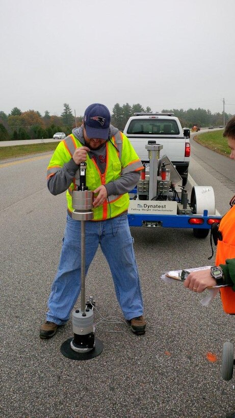 CRREL civil engineering technician Andy Bernier uses Light weight deflectometer (LWD) in support of the New Hampshire Department of Transportation (NHDOT) to evaluate a new highway pavement design and construction strategy incorporating geo-grid fabric.  Testing with the falling weight deflectometer (FWD) is shown in the background.