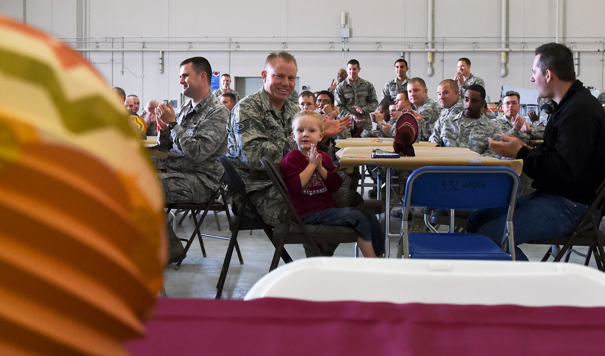 Tech. Sgt. Stephen Mills, 432nd Aircraft Maintenance Squadron Reaper support shift lead, center, and his three-year-old son Daven, applaud the opening comments at the 432nd Maintenance Group’s Thanksgiving celebration at Creech Air Force Base, Nevada, Nov. 21, 2014. Nearly 200 Airmen and family members attended the event to celebrate the holiday. (U.S. Air Force photo by Tech. Sgt. Shad Eidson/released)
