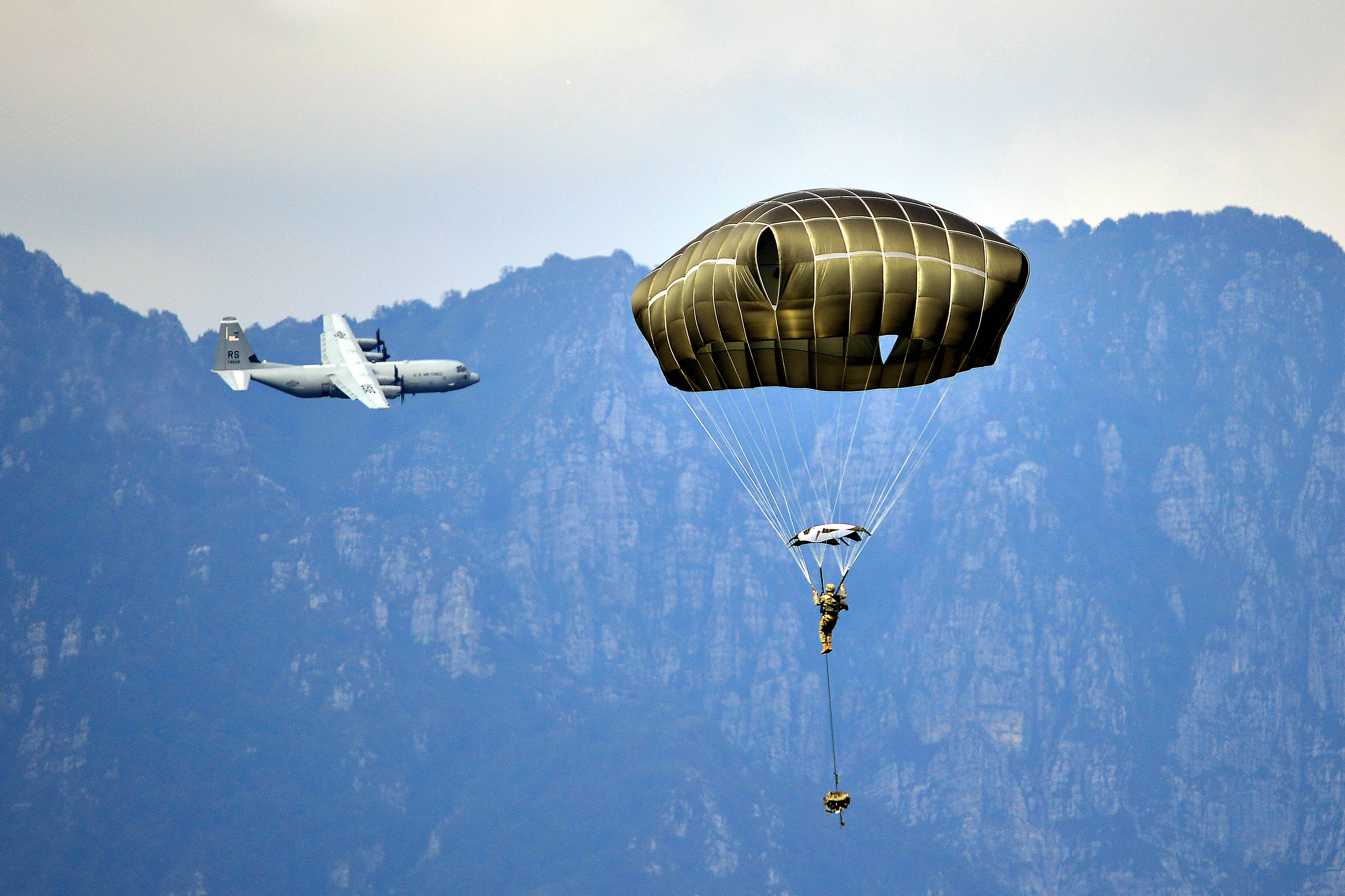 A C-130 Hercules aircraft departs while a U.S. paratrooper descends onto  the Juliet Drop Zone