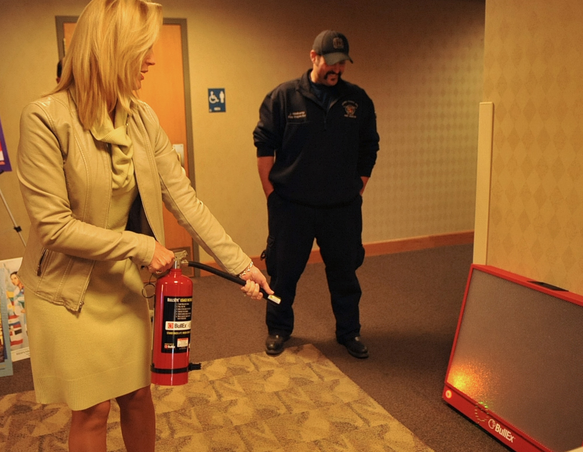 Julie McWalter, 319th Force Support Squadron marketing director, practices effectively using a portable fire extinguisher while Justin Haskamp, Grand Forks Air Force Base Fire Department fire inspector, offers instruction Oct. 8, 2014, at Grand Forks AFB, N.D. Members were able to put out a simulated fire on the BullEx Bullseye Extinguisher Training System which uses sensors to detect an infrared laser coming from the fire extinguisher to adjust the flames accordingly. The machine can sense movement to know how close or far away the individual is, and it can also be programmed to know what type of extinguisher is being used. The training offered by the Grand Forks AFB Fire Department was a part of Fire Prevention Week Oct. 5-11. (U.S. Air Force photo/Airman 1st Class Bonnie Grantham)