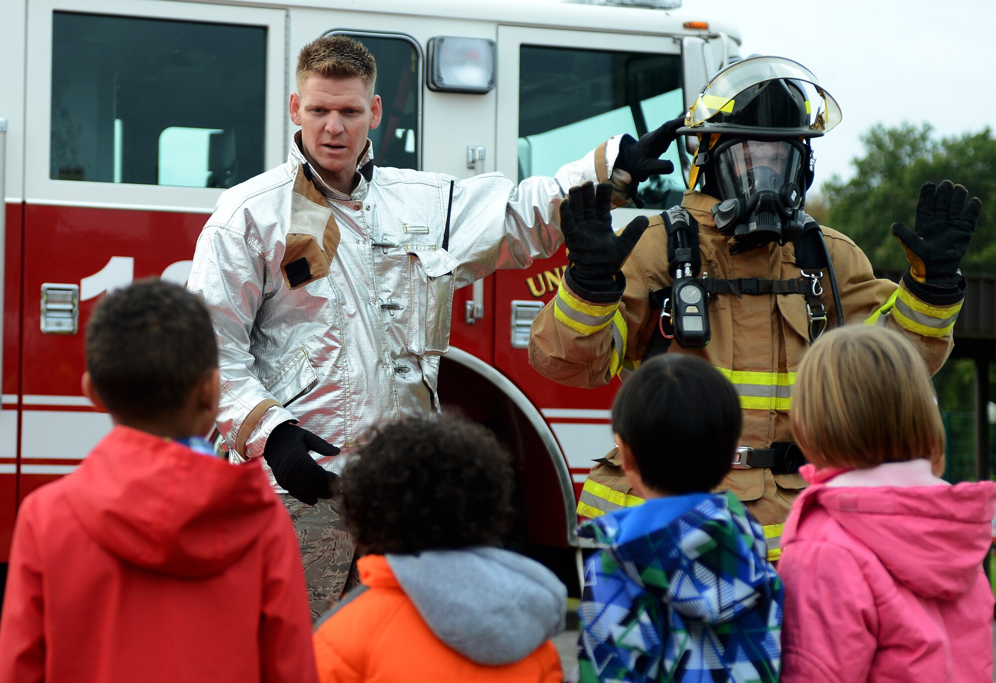 U.S. Air Force Tech. Sgt. Andrew J. Kehl, a 52nd Civil Engineer Squadron firefighter and native of Palm Springs, Calif., teams up with U.S. Air Force Airman 1st Class Daniel R. Prokowich, 52nd CES firefighter and native of Clermont, Calif., to show Spangdahlem Elementary School students firefighter equipment while visiting SES on Spangdahlem Air Base, Germany, Oct. 8, 2014. The additional gear a firefighter wears weighs approximately 75 pounds. (U.S. Air Force photo by Airman 1st Class Luke J. Kitterman/Released)