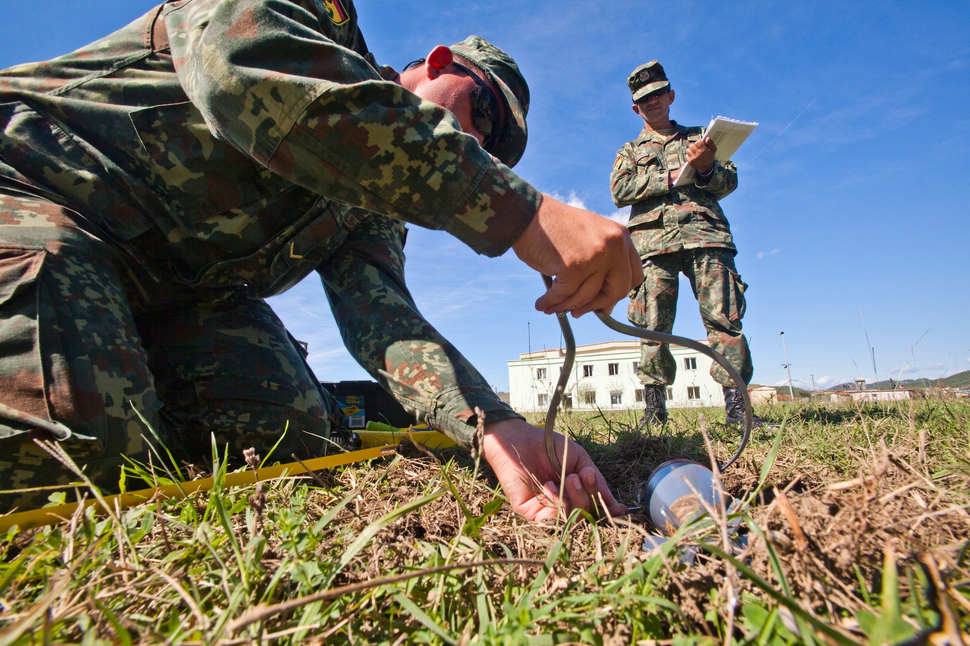 A picture of Albanian Army Cpl. Hajredin Istrefi observing Lance Cpl. Dorjan Isufi, measure an unexploded munition.