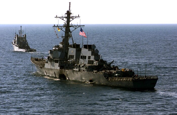 The crew of the Arleigh Burke-class USS Cole (DDG 67) escort their wounded ship aboard Navy tug vessel, USNS Catawaba, to a staging point in the Yemeni harbor of Aden awaiting transportation by the Norwegian-owned semi-submersible heavy lift ship MV Blue Marlin back to their homeport, during Operation Determined Response, on Oct. 29, 2000.