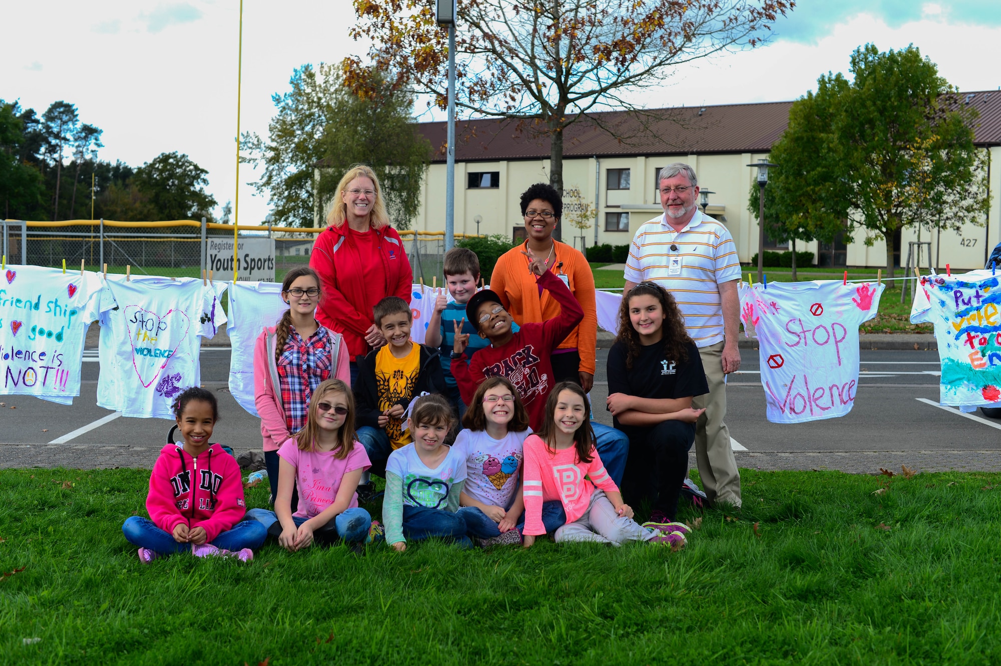 Karen Pilalas, 4th-grade child and youth programs assistant at the 52nd Force Support Squadron School Age Program, poses with her class in front of the School Age building on Spangdahlem Air Base, Germany, Oct. 14, 2014. The clothes line displaying painted shirts brings awareness to domestic violence and is hung behind the class. 52nd Medical Operations Squadron Family Advocacy hosted the shirt hanging at the School Age Program facility to help encourage children to recognize domestic violence and report it to the authorities. (U.S. Air Force photo by Airman 1st Class Kyle Gese/Released)