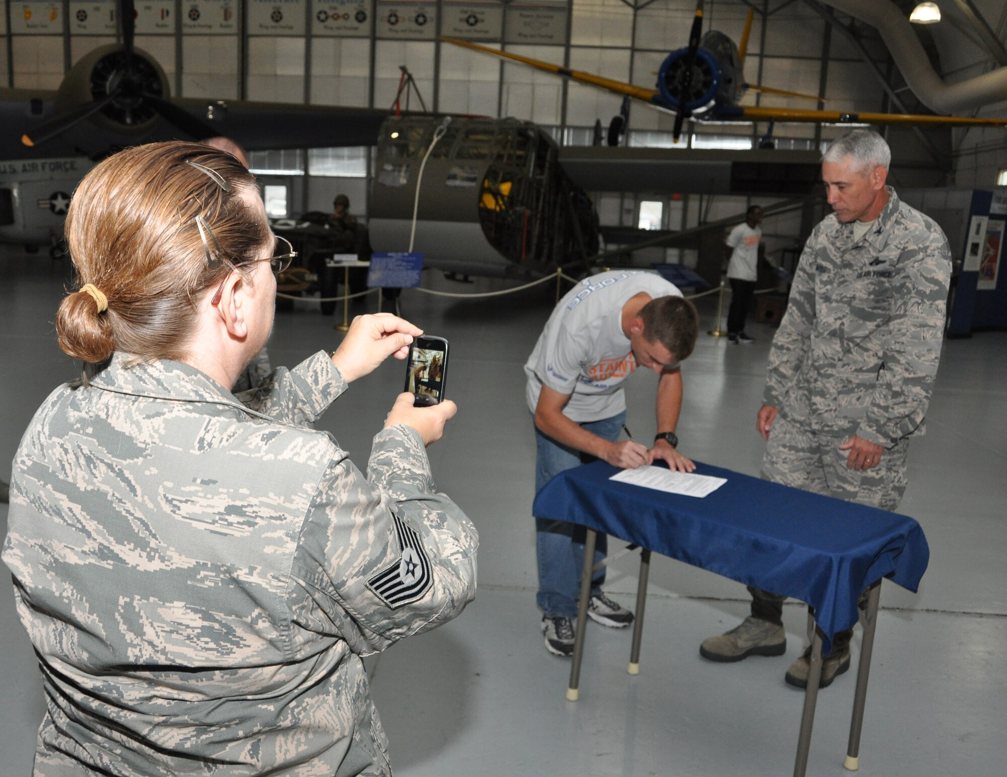 Tech. Sgt. Melena Quetel (left), 46th Aerial Port Squadron, takes a photo of her son, Justin Quetel (middle), as he signs an enlistment contract to serve in the Air Force Reserve Oct. 4, 2014, at the Air Mobility Command Museum, Dover, Del. Col. J. Scott Hayes, 512th AW vice commander administered the oath of enlistment to Quetel and five others as part of a mass enlistment ceremony attended by family and friends. One week prior, Quetel completed his service to the U.S. Marine Corps, after serving eight years. (U.S. Air Force photo/Senior Airman Joe Yanik)