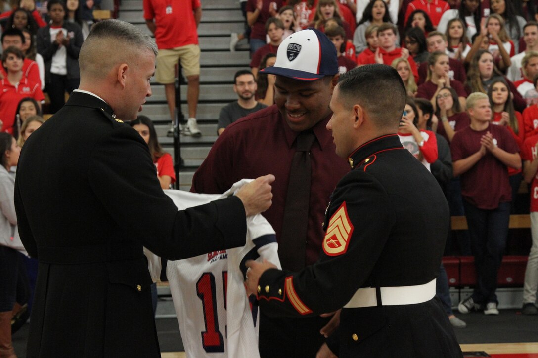 Malik McMorris (center), a defensive lineman and fullback for the Mater Dei High School Monarchs and 17-year-old native of Santa Ana, Calif., receives his Semper Fidelis All-American Bowl jersey from Maj. James P. Dollard (left), the commanding officer of Marine Corps Recruiting Station Orange County, and Staff Sgt. Jeno Jorgensen, a local Marine recruiter, during a pep rally at Mater Dei High School in Santa Ana, Oct. 17, 2014. McMorris is among approximately 100 student athletes from across the nation selected to participate in the game at the StubHub Center in Carson, Calif., Jan. 4, 2015. He is currently the only 2015 selectees from Orange County.