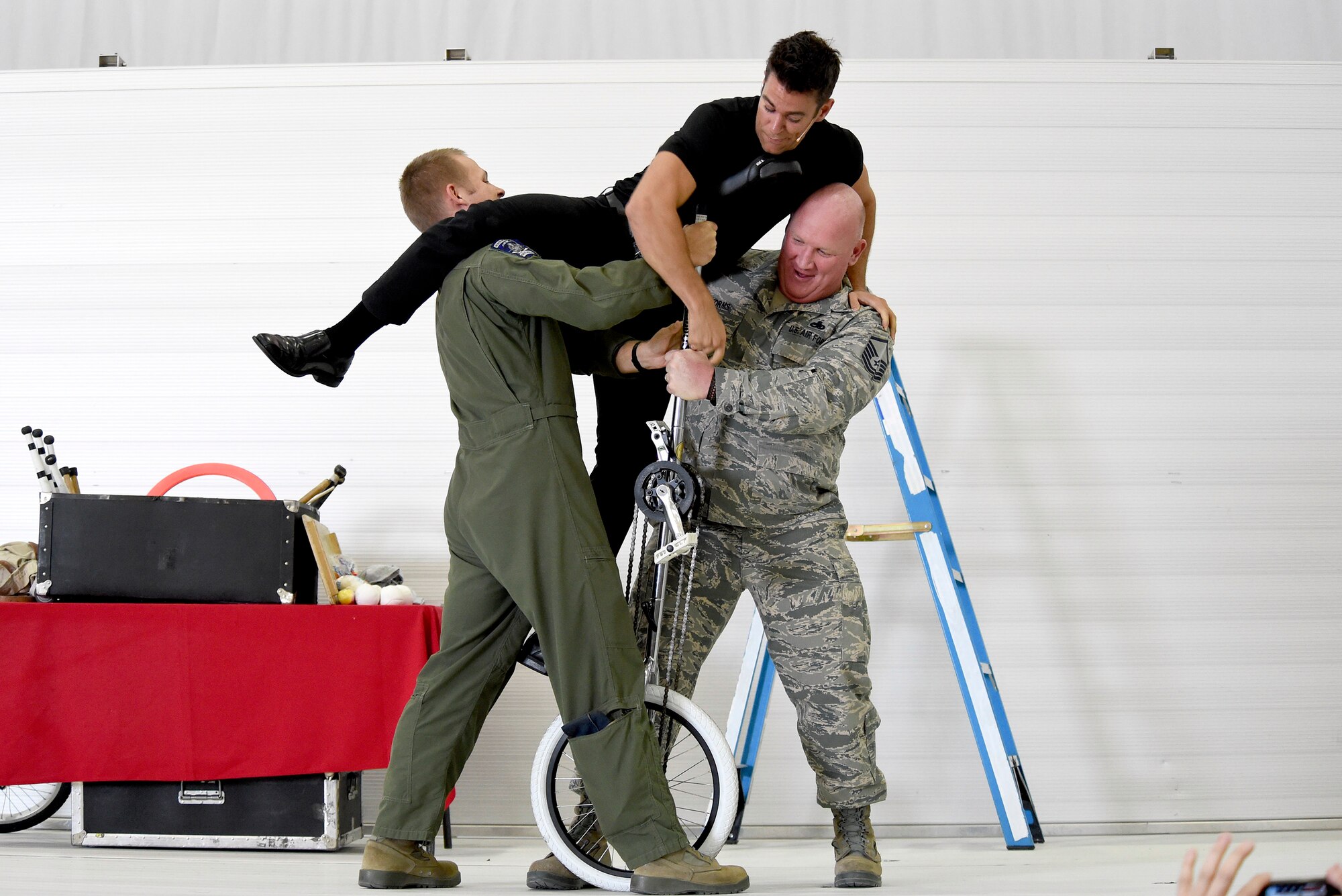 Capt. Travis, left, 42nd Attack Squadron director of staff, and Master Sgt. Dustin Storms, right, 432nd Aircraft Maintenance Squadron Programs Section NCO in charge, help juggler and comedian, Jeff Civillico, climb onto a unicycle during his performance at Creech Air Force Base, Nevada, Oct. 17, 2014. The entertainment and food event was the latest USO-sponsored show to lift the spirits of America’s troops and their families, which the USO has provided since its creation in 1941. (Last names have been withheld for security purposes). (U.S. Air Force photo by Tech. Sgt. Shad Eidson/Released) 