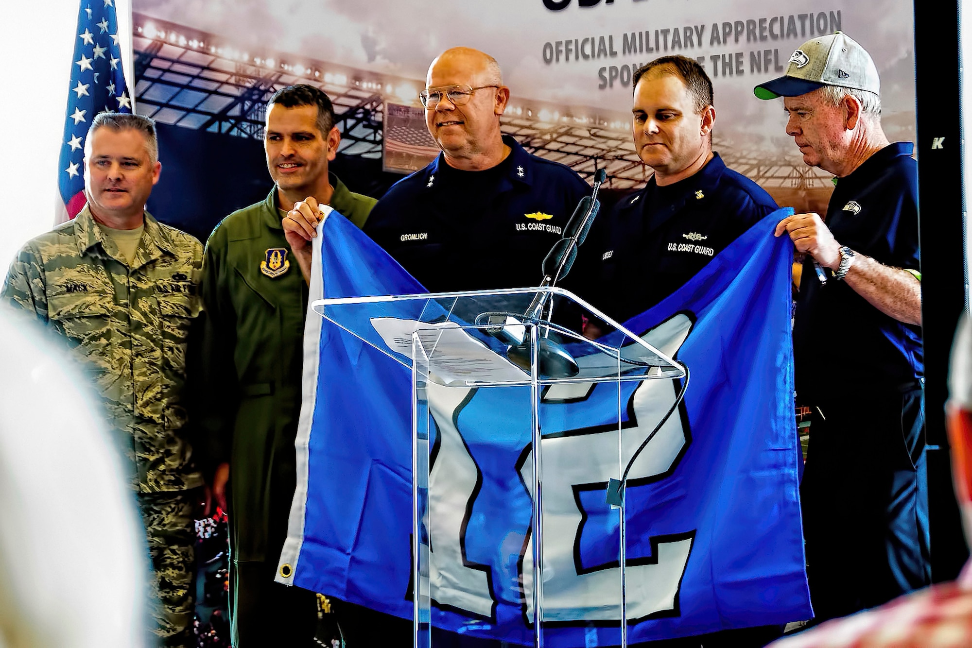 On Sept. 9, the 446th Airlift Wing's Command Chief Master Sgt. Tony Mack (left to right) and Col. Rick Grayson, commander, handed over the reins as the Seattle Seahawks championship football team's military unit for the season to the Coast Guard District 13 Seattle. Accepting the 2014 season Seahawks military unit honors are Rear Adm. Richard Gromlich  and Command Master Chief Charles Lindsey Coast Guard District 13.  Far right, they are joined on stage by Mike Flood, vice president of community affairs for the Seattle Seahawks. (Air Force Reserve photo by Jake Chappelle)