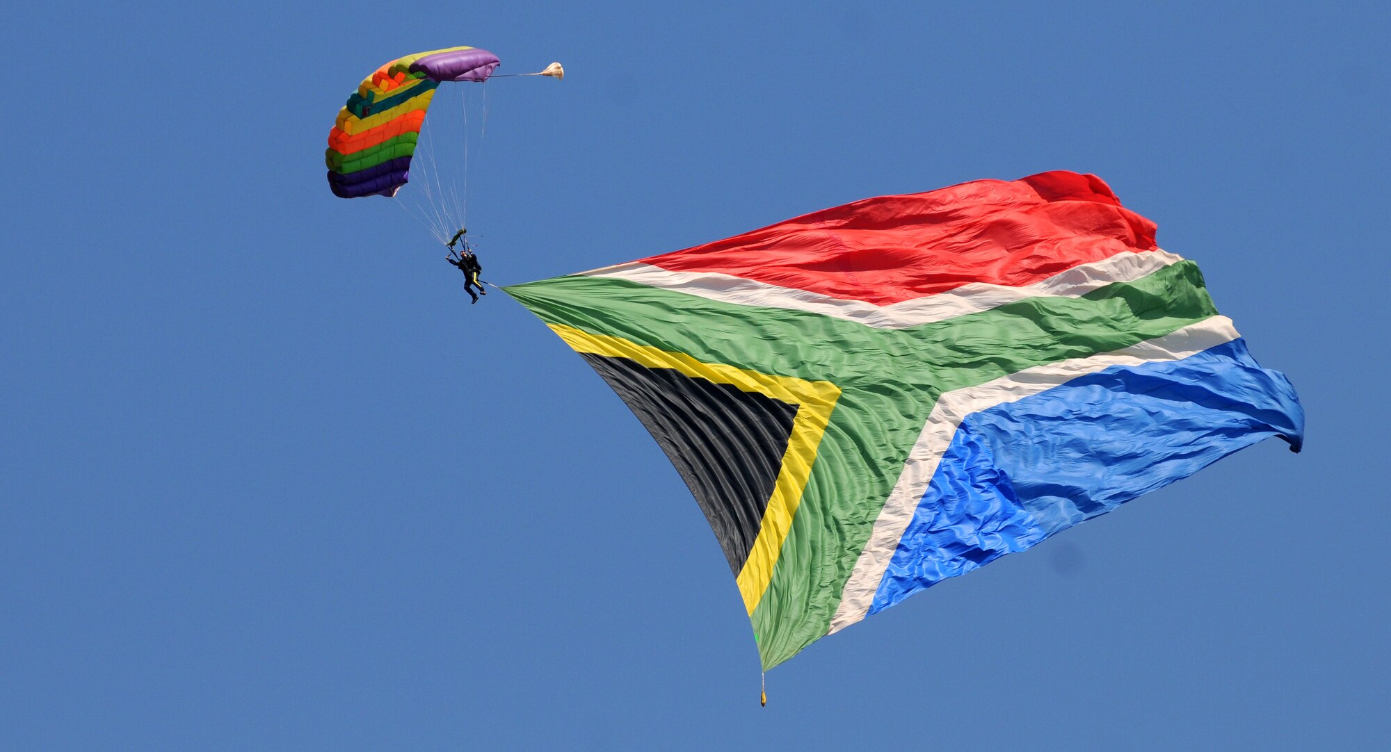 A parachutist descends into the opening ceremony of the Africa Aerospace & Defence Expo 2014 Sept. 17, 2014, at Waterkloof Air Force Base, Pretoria, South Africa, towing the South African national flag. U.S. Air Force Airmen and U.S. Army Soldiers attended the opening day of the 8th annual multinational event. United States military aircraft, including a C-130J Super Hercules and C-17 Globemaster III were on static display at the show, and the U.S. Army Golden Knights parachute demonstration team jumped into the opening ceremony alongside their South African counterparts, the Golden Eagles. (U.S. Air Force photo by Tech. Sgt. Austin M. May/Released)