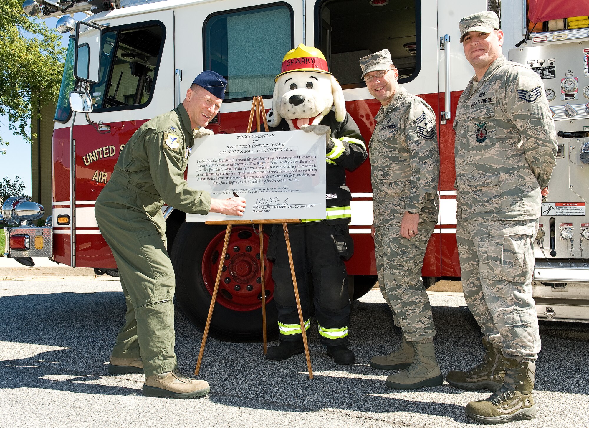 Col. Michael Grismer, 436th Airlift Wing commander, left, signs the 2014 Fire Prevention Week proclamation Sept. 26, 2014, on Dover Air Force Base, Del. Sparky the fire dog, Chief Master Sgt. Stanley Cadell, 436th AW command chief, and Staff Sgt. Matthew Woods, 436th Civil Engineer Squadron fire inspector, witnessed the signing of the proclamation stating Oct. 5-11 as fire prevention week at Dover AFB and delivering a campaign message of "Working smoke alarms save lives: test yours every month!" (U.S. Air Force photo/Roland Balik)