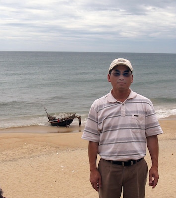 Karl Mai stands on the same beach from which he left Vietnam in 1981 as one of the fleeing “Boat People.” Behind him is a bamboo fishing boat like the one he and 18 others crowded into for the nighttime escape to Hong Kong. This photo was taken in August 2012 when Mai and his family revisited Vietnam.