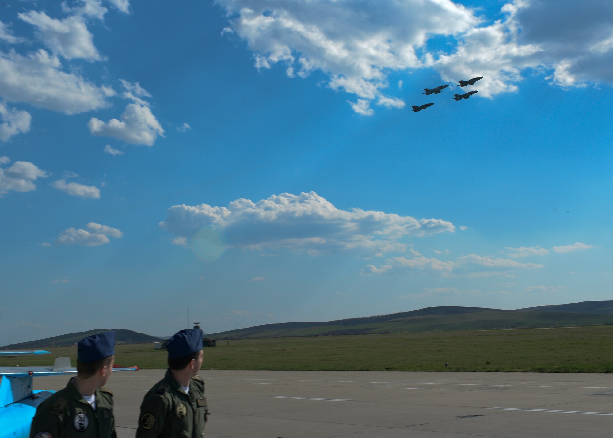 Two Romanian air force pilots watch as a formation of four Romanian air force MiG-21 fighter aircraft fly above the flightline at Campia Turzii, Romania, April 16, 2015. The U.S. and Romanian air forces will conduct training aimed to strengthen interoperability and demonstrate the countries' shared commitment to the security and stability of Europe. (U.S. Air Force photo by Staff Sgt. Joe W. McFadden/Released)