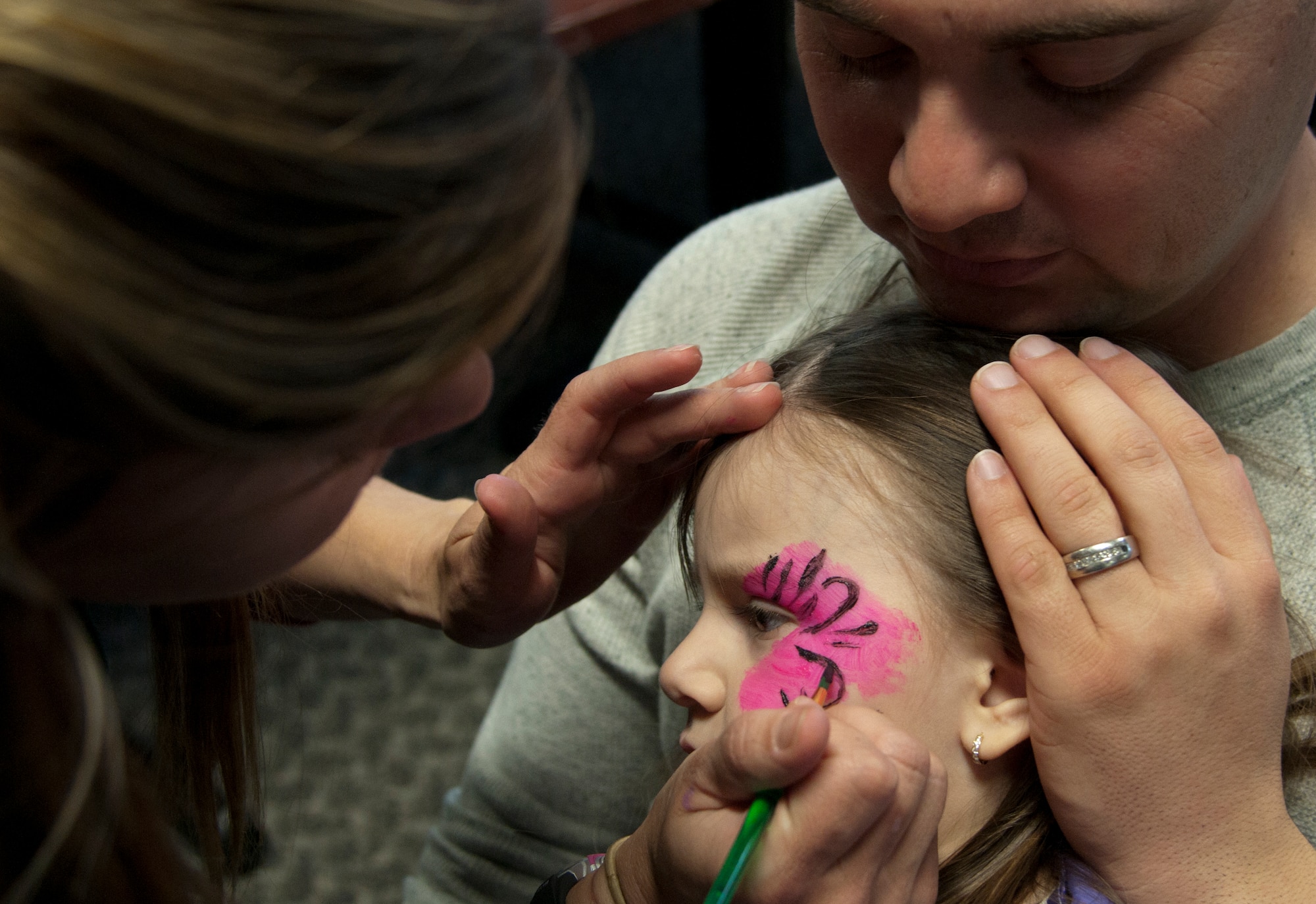 Amy Dent, 90th Force Support Squadron, paints a flower onto Alyssa Avila, while her father, Staff Sgt. Johnathan Avila, 90th Civil Engineer Squadron, holds her April 17, 2015, during the Spring Carnival in the Fall Hall Community Center, F.E. Warren Air Force Base, Wyo., April 17, 2015. Children were offered a multitude of designs to choose from, including superheroes, flowers and (U.S. Air Force photo by Airman 1st Class Brandon Valle)