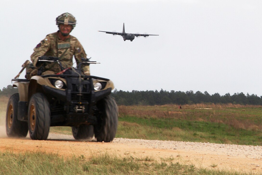 A British paratrooper rides a four-wheeled vehicle while a British air force C-130 Hercules aircraft approaches for landing on Fort Bragg, N.C., April 14, 2015.