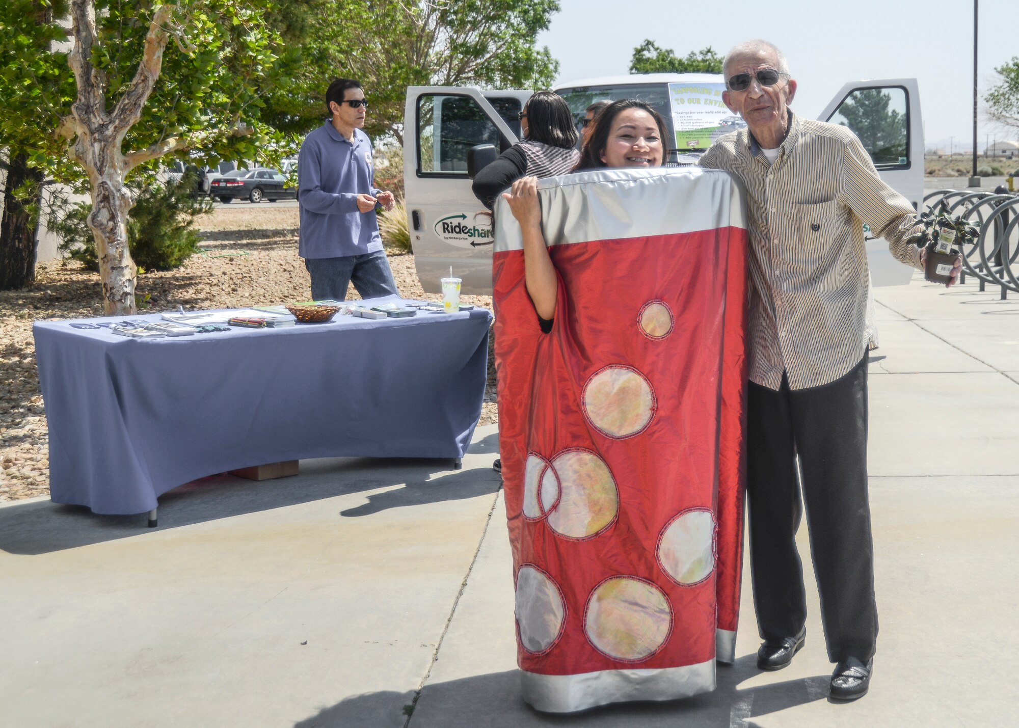 MyNga Day, environmental planner, dressed up as an aluminum can to encourage people to recycle. With her, Robert Patete, retired sailor, shows off the plant he won from Corvias Military Housing during the Earth Day celebration April 22 at the Exchange. Behind them, a Ride Share van reminds base personnel to consider carpooling to work to conserve resources and keep the air cleaner. (U.S. Air Force photo by Rebecca Amber)
