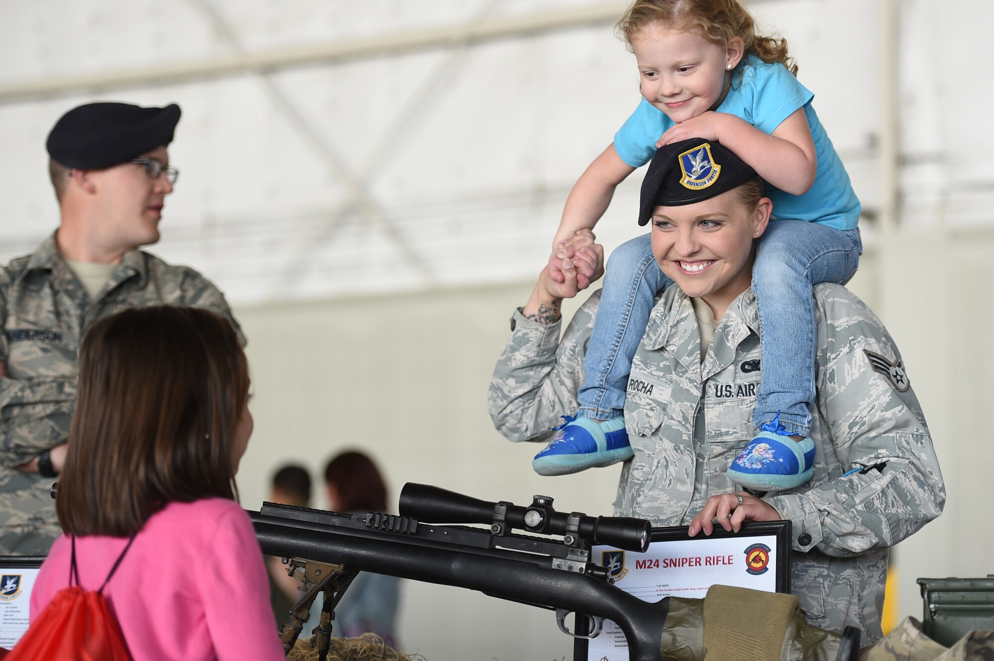 Senior Airman Stephanie Rocha, 799th Security Forces Squadron combat arms training and management  instructor, carries her daughter, Christina, three, while she explains the capabilities of an M24 Sniper rifle at the “Take Your Kid to Work Day” event April 23, 2015 at Creech Air Force Base, Nevada. Creech families honored the Month of the Military Child with food, face painting, and a magic show sponsored by the Las Vegas USO. In addition military vehicles and equipment were also on display and included the MQ-1 Predator and MQ-9 Reaper aircraft.  (U.S. Air Force photo by Tech Sgt. Nadine Barclay/Released)