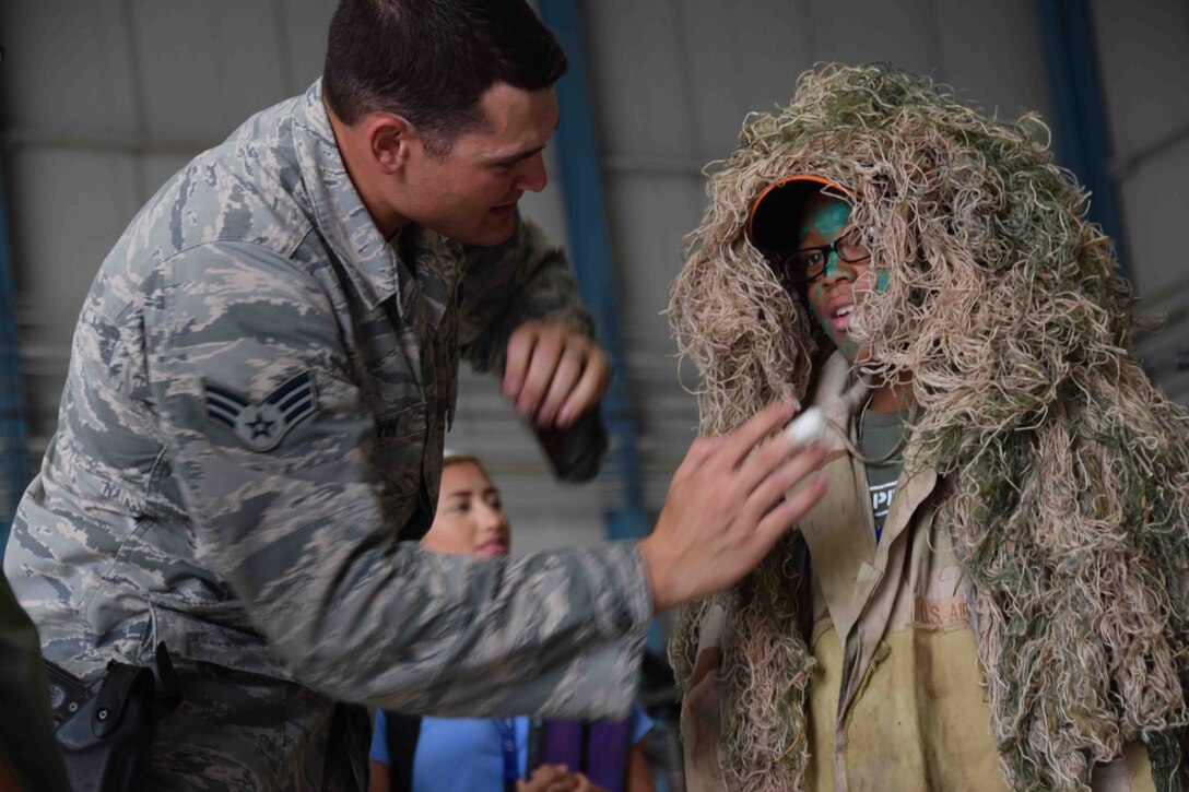 Combat weapons instructor, Senior Airman Cory Irwin from the 37th Training Squadron, helps Raphael Webb don a Ghillie suit, at the weapons display at Joint Base San Antonio-Lackland’s Operation Junior Expeditionary Team on July 31, 2015. The children also climbed aboard a Humvee and toured a C-5A Galaxy aircraft. (U.S. Air Force photo/Tech. Sgt. Carlos J. Trevino)