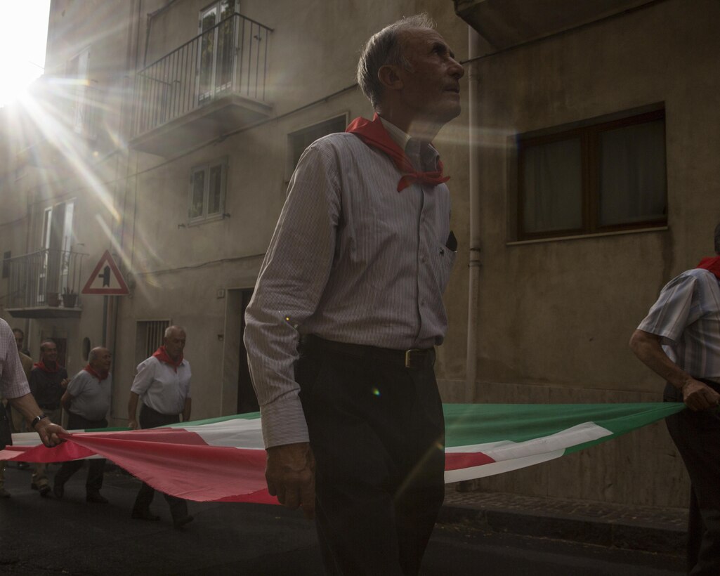 An Italian military veteran leads a parade with Marines and sailors with Special-Purpose Marine Air-Ground Task Force Crisis Response-Africa, Detachment A, through the street carrying the Italian flag August 2, 2015. The Marines and sailors also observed a mass at the Catherdral of Maria SS Annunziata and participated in a wreath-laying ceremony in commemoration of the Battle of Troina with Italian civil and military representatives as well as the people of Troina. (U.S. Marine Corps photo by Cpl. Olivia McDonald/Released)
