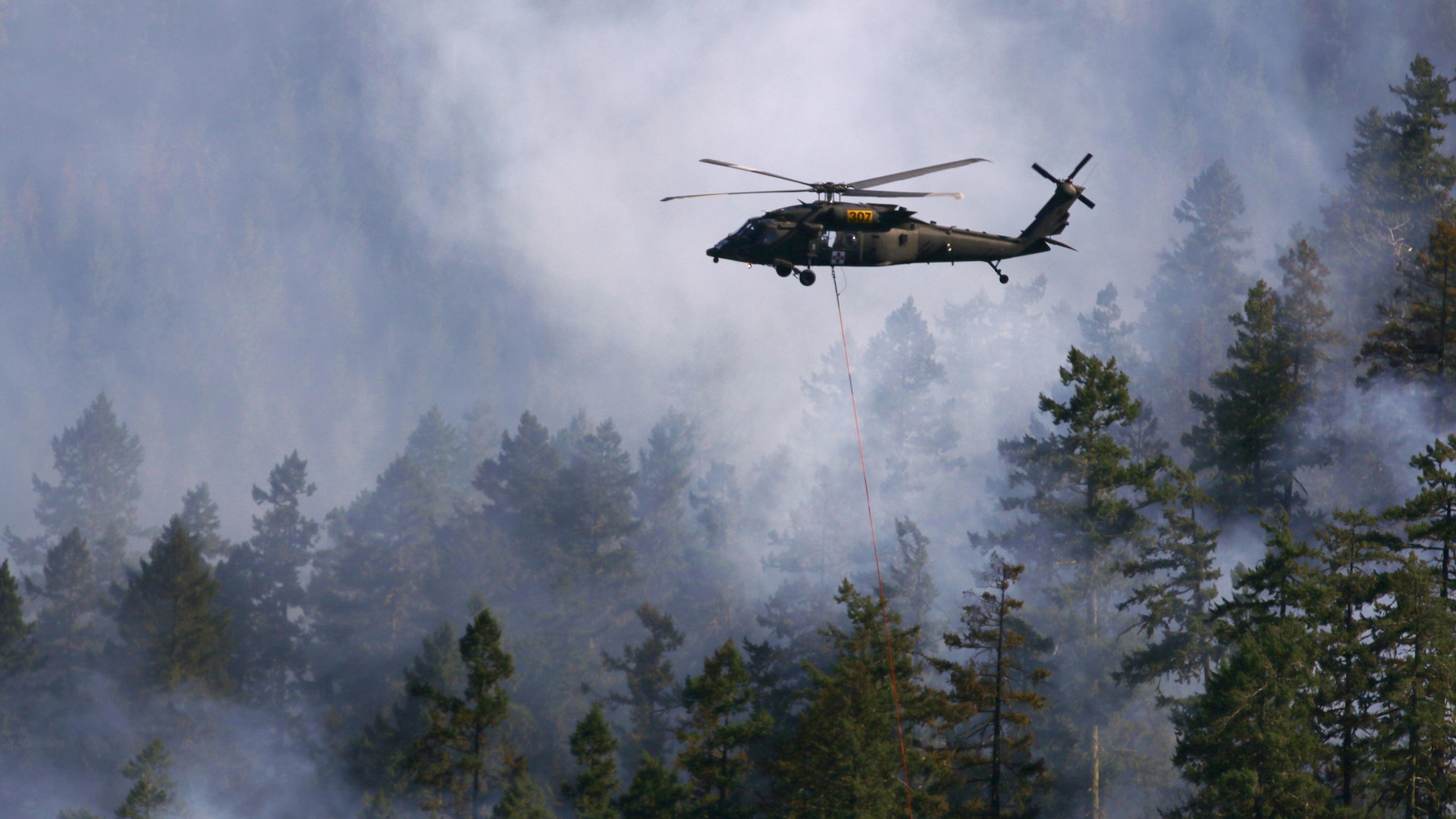 Oregon Army National Guard pilots, Chief Warrant Officer 4 Dennis Cooper, Chief Warrant Officer 2 Logan Bass, and crew chief Spc. Beth Bechard, with Charlie Company, 7-158th Aviation, navigate through smoke on the way to their drop site in support of firefighting ground crews, Aug. 5, at the Stouts Fire. The Black Hawk helicopter is equipped with a "Bambi bucket" which carries approximately 500 gallons of water. Two Oregon Army National Guard HH-60M Black Hawk helicopters have been working out of the Roseburg Municipal Airport since Monday to support ground firefighting crews and have been joined by one Oregon Army National Guard CH-47 Chinook helicopter based out Pendleton, Oregon.