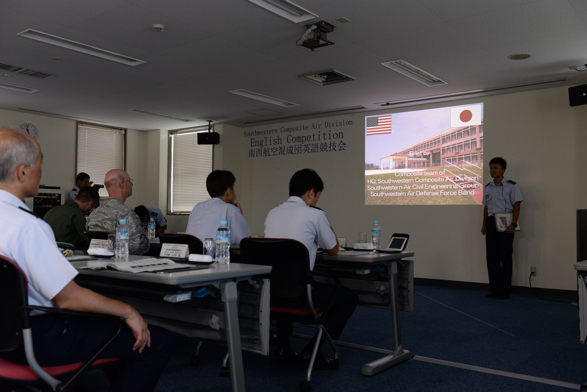 Japan Air Self Defense Force Airman gives a briefing in English as United States Air Force Maj. Christopher L. Teke, 18th Civil Engineer Squadron, and Maj. Mark Carl, 909th Air Refueling Squadron, judge how well they presented at Naha Air Base, Japan, Aug. 26, 2015. JASDF hosted an English briefing contest to be judged by American Airmen. (U.S. Air Force photo by Senior Airman Omari Bernard)