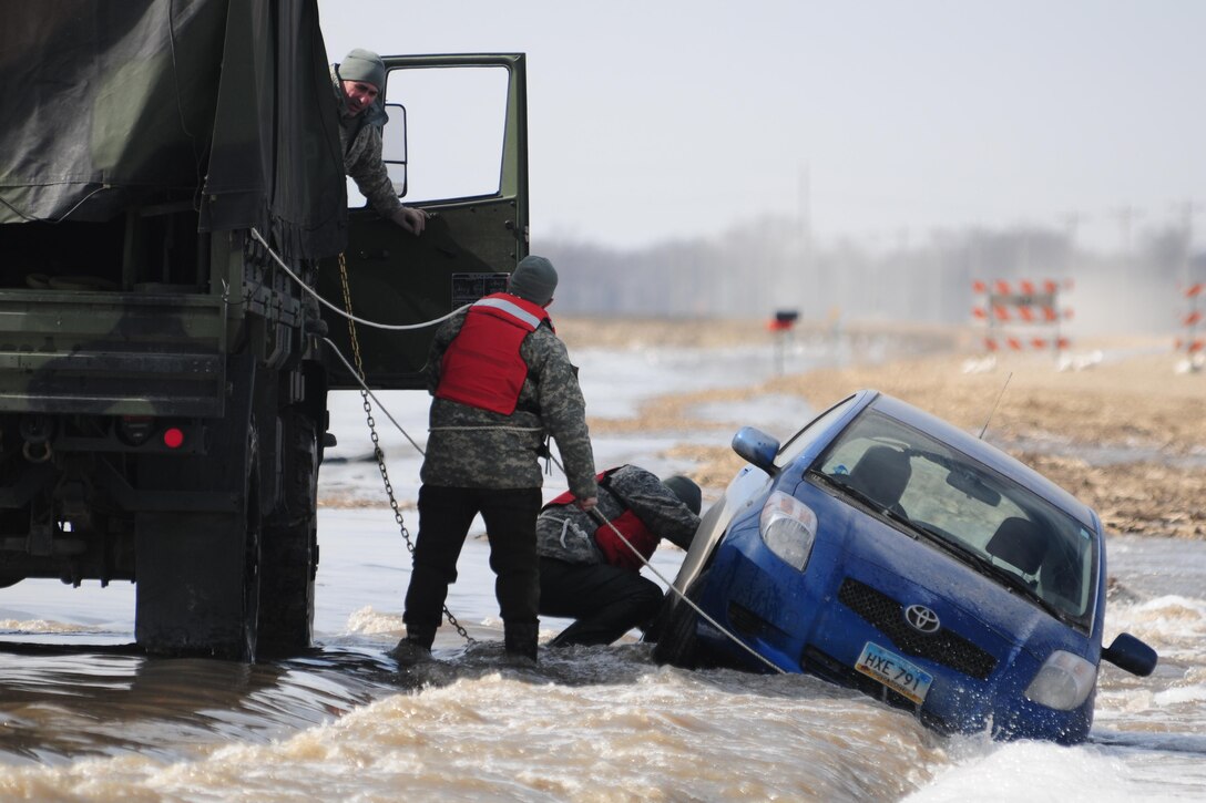 North Dakota Army National Guardsmen Sgt. Preston Steele, right, and Spc. Jeremy Kasperson attach a towing chain to a partially submerged vehicle on a washed out portion of a gravel road a few miles west of Harwood, N.D., March 25, 2015. The driver of the vehicle was able to escape the car and make his way to safety before help arrived. The soldiers are assigned to the 815th Engineer Company. North Dakota Air National Guard photo by Senior Master Sgt. David Lipp