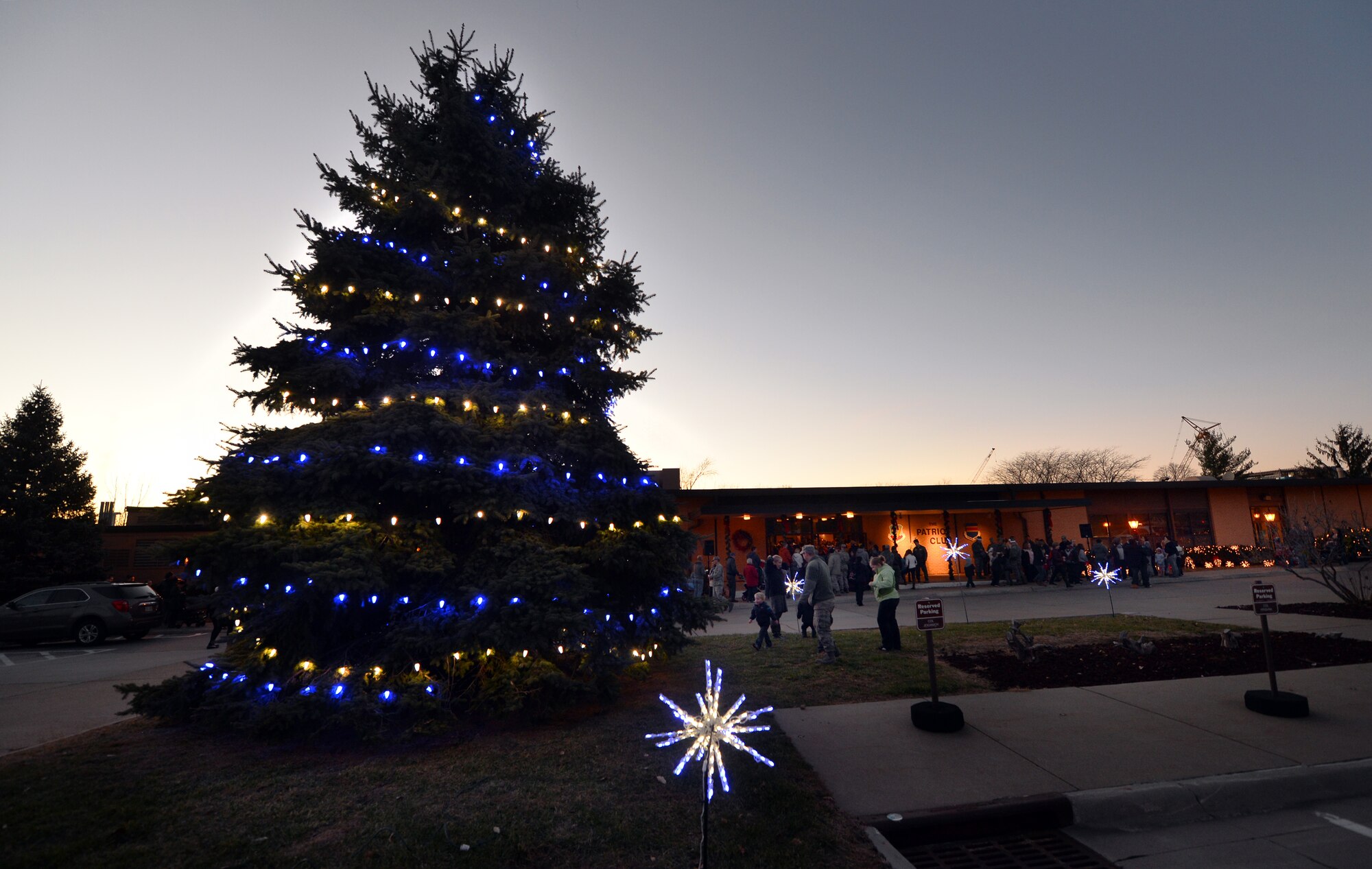 The official tree for the annual Tree Lighting Ceremony comes to life with strings of blue and white lights just outside of the Patriot Club on Dec. 3, Offutt Air Force Base, Neb.  Lines of people wait their turn to enter the club.  The ceremony hosts a full buffet, photos with Santa and Mrs. Claus, numerous vendors, distinguished visitors, live music and endless hot chocolate and sweets.  (U.S. Air Force photo by Josh Plueger/Released)