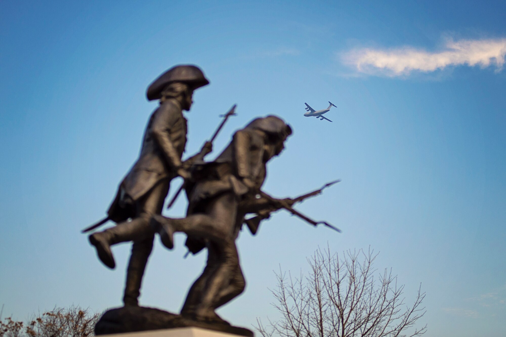 A Team Dover C-5M Super Galaxy flies above after takeoff with the Delaware Continentals memorial in the foreground, taken December 7, 2015, in downtown Dover, Del. Dec 7 marks the anniversary of the Pearl Harbor attacks and also Delaware Day, a state holiday to recognize the state being the first to ratify the Federal Constitution in 1787. (U.S. Air Force photo/Capt. Bernie Kale)