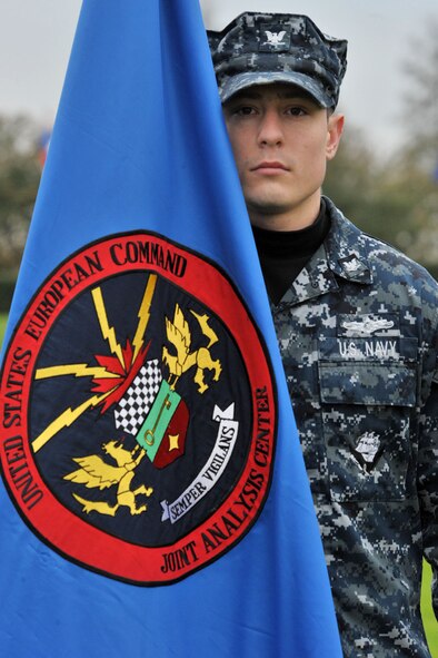 U.S. Navy Petty Officer 3rd Class Austin, Joint Intelligence Operations Center Europe Analytic Center division analyst, stands at attention with the JAC flag during the 74th Pearl Harbor Commemoration Ceremony at RAF Molesworth, United Kingdom, Dec. 7, 2015. The ceremony included a three-volley salute by the 423rd Honor Guard and a formation salute as the 48th Fighter Wing F-15 Eagle pilots performed a flyby. (U.S. Air Force photo by Staff Sgt. Ashley Tyler/Released)(Last name removed for security purposes)