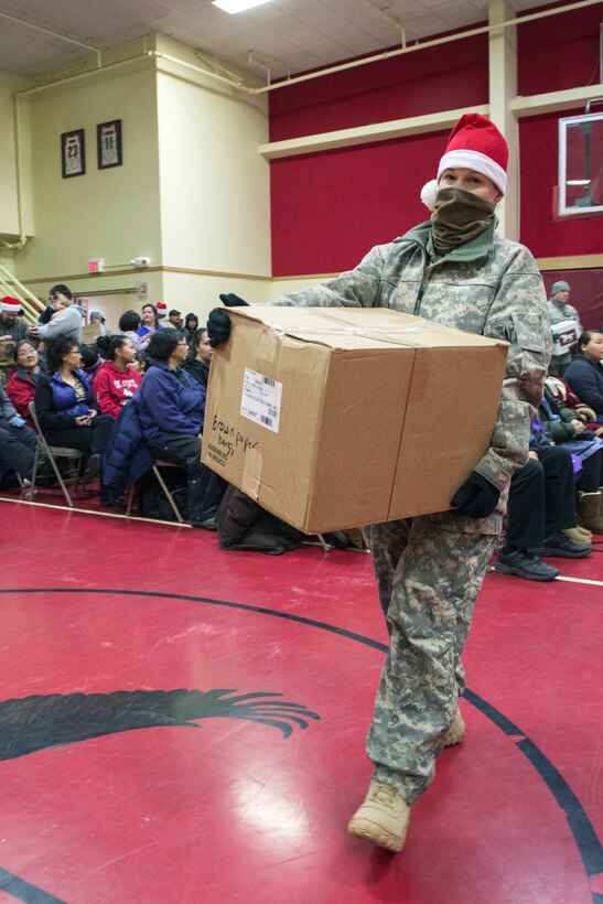 A service member wearing a Christmas hat carries a box of presents to be given to underserved children during an Operation Santa Claus delivery to the village of St. Mary's, Alaska, Dec. 5, 2015. U.S. Air Force photo by Alejandro Pena