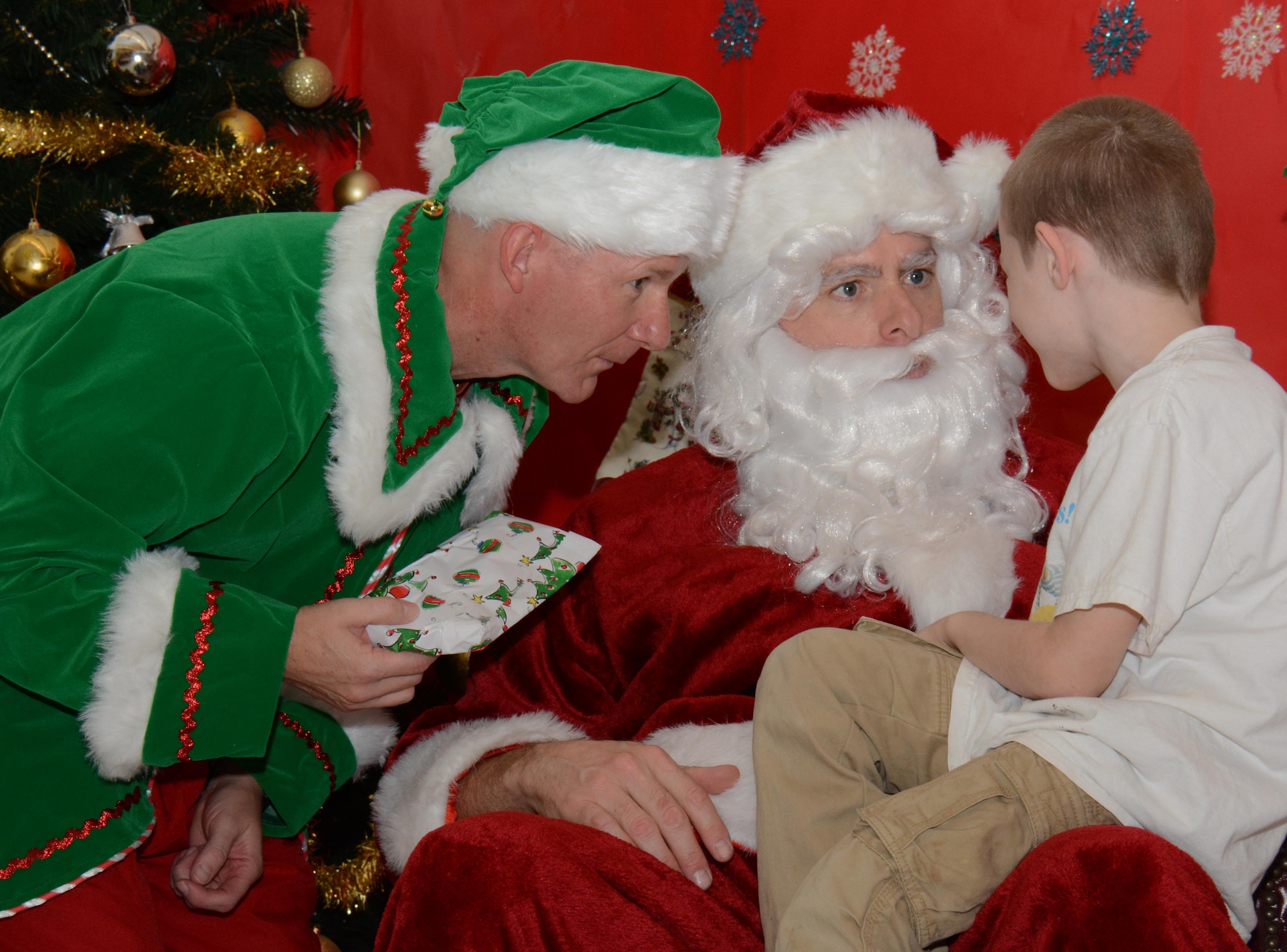 Chief Master Sgt. Thomas Good (left), 379th Air Expeditionary Wing command chief, holds a present for a child attending the Breakfast with Santa event and listens as the child explains what he wants for Christmas to Santa, Brig. Gen. Darren James (center), 379 AEW commander, inside the Blatchford-Preston Complex Dining Facility at Al Udeid Air Base, Qatar Dec. 19. Nearly 20 families took part in the event which featured a pajama contest, designing holiday stockings, decorating gingerbread cookies and coloring. (U.S. Air Force photo by Tech. Sgt. James Hodgman/Released)