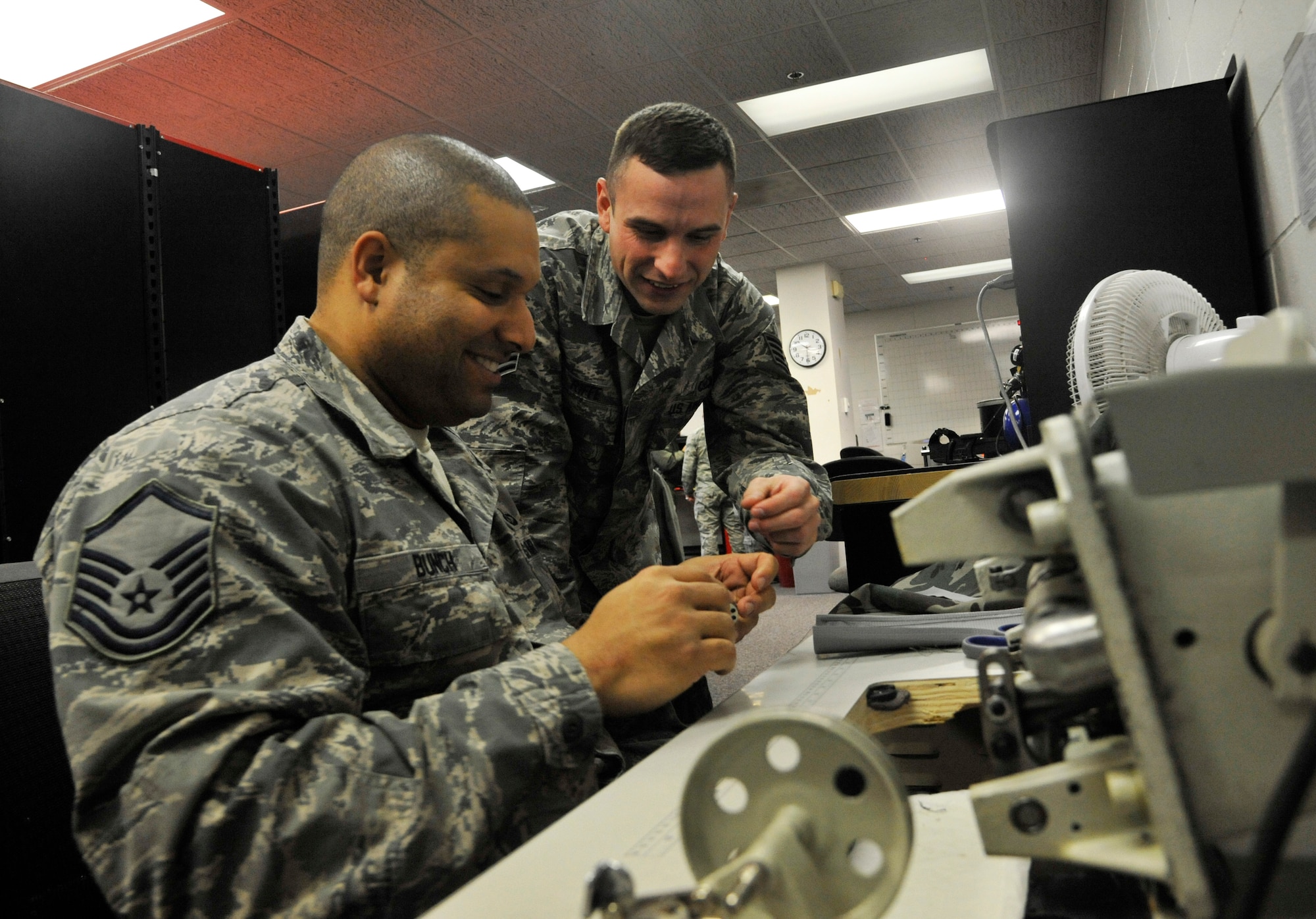 Staff Sgt. Riffe, 509th Operation Support Squadron aircrew flight equipment supervisor, assists Master Sgt. Damien Bunch, 509th OSS AFE craftsman, with a sewing machine at Whiteman Air Force Base, Mo. Jan. 11, 2015. Riffe enjoys joking while assisting others to alleviate potential frustration with the challenges at work. (U.S. Air Force photo by Senior Airman Keenan Berry/Released)