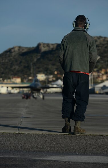 U.S. Air Force Airman 1st Class Zachary Jung, an assistant crew chief assigned to the 52nd Expeditionary Aircraft Maintenance Squadron, observes as an F-16 Fighting Falcon fighter aircraft pilot assigned to the 480th Expeditionary Fighter Squadron taxis on the flightline during a flying training deployment at Souda Bay, Greece, Jan. 30, 2015. Jung used a series of hand gestures and movements to convey to the pilot whether to stop or keep moving while the aircraft taxied on the flightline. (U.S. Air Force photo by Staff Sgt. Joe W. McFadden/Released)