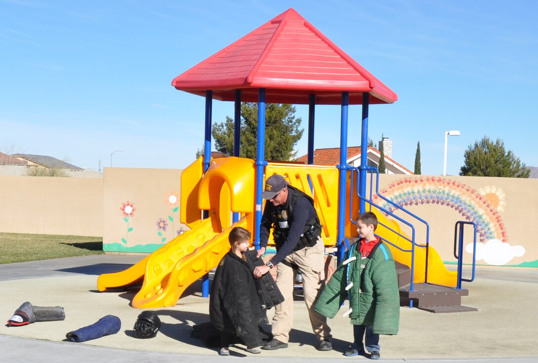 Sgt. Steven Goss, K-9 trainer aboard Marine Corps Logistics Base Barstow assists Ryley and Logan Witcher, the five-year-old daughter and seven-year-old son of Lt. Justin Witcher, accident investigations chief at MCLB Barstow, to try on parts of a suit used for K-9 training during a demonstration at Endeavor Elementary School, Feb. 4.  Ryley and Logan are students at EES and Logan’s presentation regarding K-9 officers inspired the demonstration
