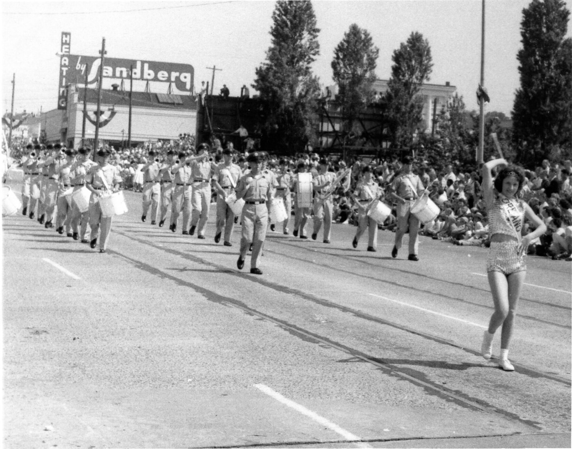 The OreANG’s Twirler, probably champion Ms. Carol Johnson, leads the OreANG Drum & Bugle Corps in the summer khaki uniforms of that era down a Portland street in the 53rd Annual Rose Festival’s Grand Floral Parade, Saturday, June 10, 1961. Drum Major MSgt Unverricht leads the corps immediately behind her.
