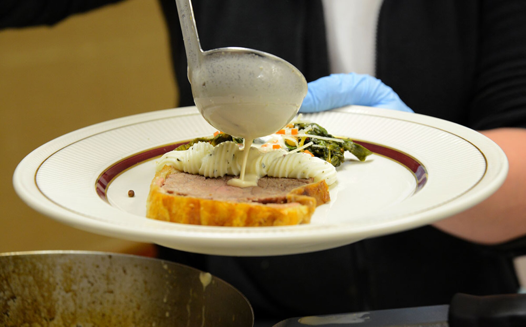 The green peppercorn sauce is poured carefully over the main course; a Beef Wellington, a side of herbed mashed potatoes and warm wilted green salad, served at the Kenai Dining Hall, JBER on Feb. 20, 2015. The 10-person team of Soldiers and Airmen tried to make a five-star three-course meal from an Army field kitchen for JBER leadership to prepare for 40th Annual Military Culinary Arts Competitive Training Event at Fort Lee, VA. March 4-13. (Air Force photo/Airman Christopher R. Morales)