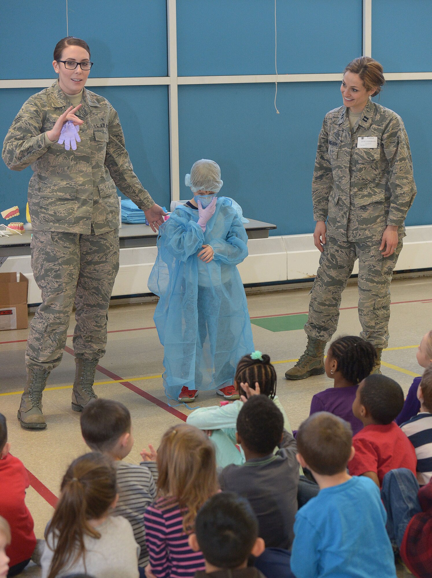 Senior Airman Jennifer Grier, dental technician, 66th Medical Squadron, describes the protective clothing a dentist would wear during an exam to kindergarteners as their classmate Aiden Heber models the clothing and Dr. (Capt.) Julie Anderson, dentist, 66th MDS, watches, during an assembly at the Hanscom Primary School, Feb. 25.  February is National Children’s Dental Health Month and programs such as this one are designed to help children understand how to take care of their teeth. (U.S. Air Force photo by Jerry Saslav)