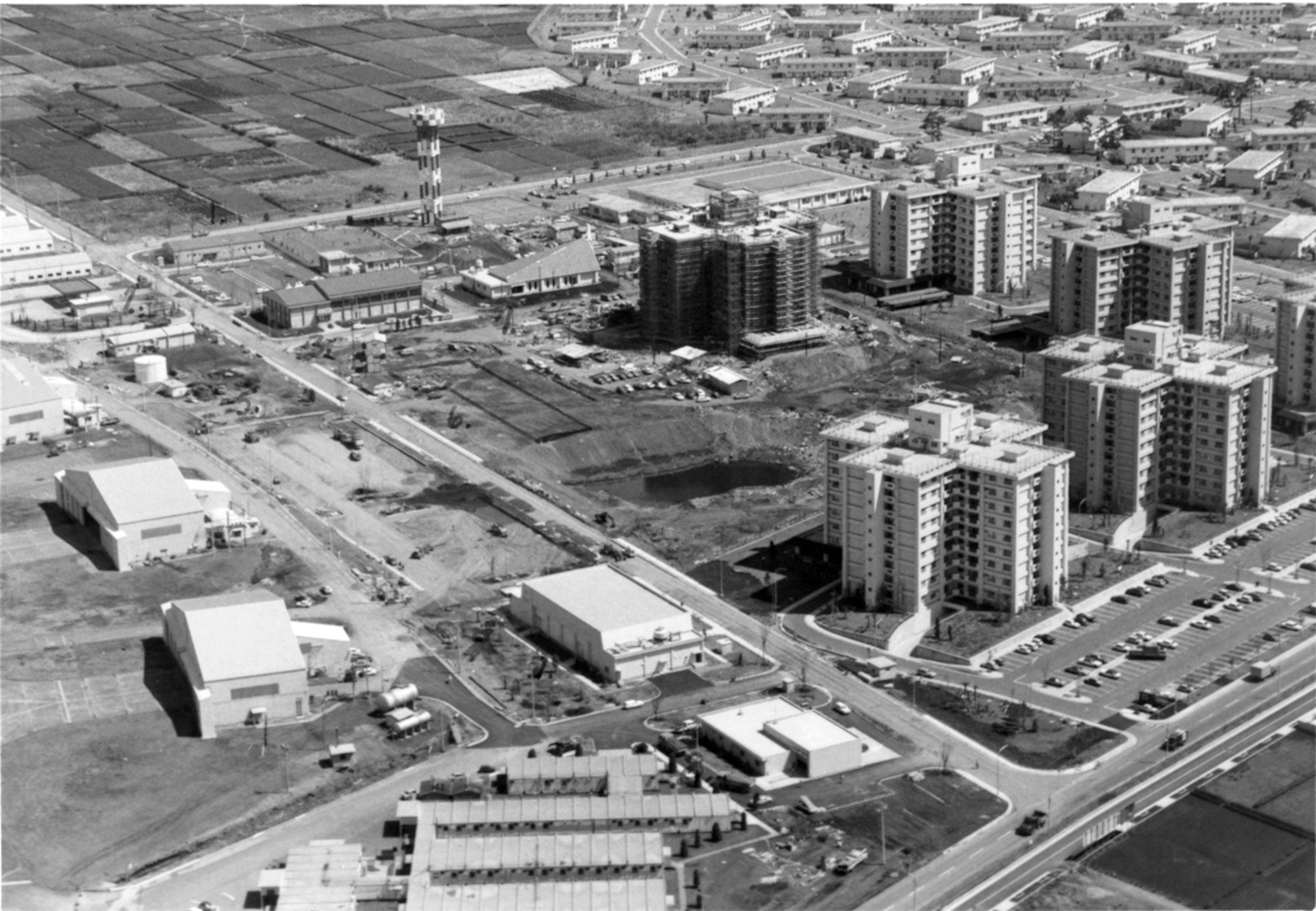 A 19 April 1977 aerial photo of Yokota's east side depicting the scope of
KPCP development. Apart from the hangars and small buildings center-left,
and the metal building at the bottom, all of the structures in this view
were newly built under KPCP. The tower apartments, East Chapel, East
Elementary School (now Mendel Elementary), movie theater, fire station,
water tower, gym, and garden housing units. Even the roads and parking lots
were new. The body of water in the center, today the location of a park, was
an old sump used for rainwater runoff until being filled in soon after this
photo was taken. (Photo courtesy of the 374th 
Airlift Wing History Office)