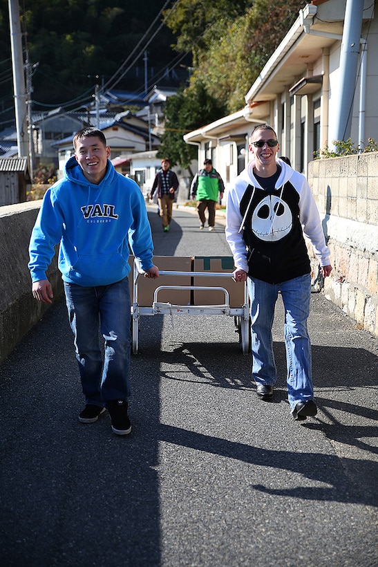 Cpl. Matthew Ahn, left, a chemical, biological, radiological and nuclear defense specialist aboard Marine Corps Air Station Iwakuni, and Cpl. Ruben Salinas, right, resident Marine Air Ground Task Force planner for Marine Aircraft Group 12, walk with their boxes of canned food to the Akebono Orphanage in Nasake Sima, Yamaguti, Japan, Dec. 23, 2014. As a part of giving back to the Japanese locals, MAG-12 Marines collected 777 cans of food from their unit holiday party to distribute to those in need.