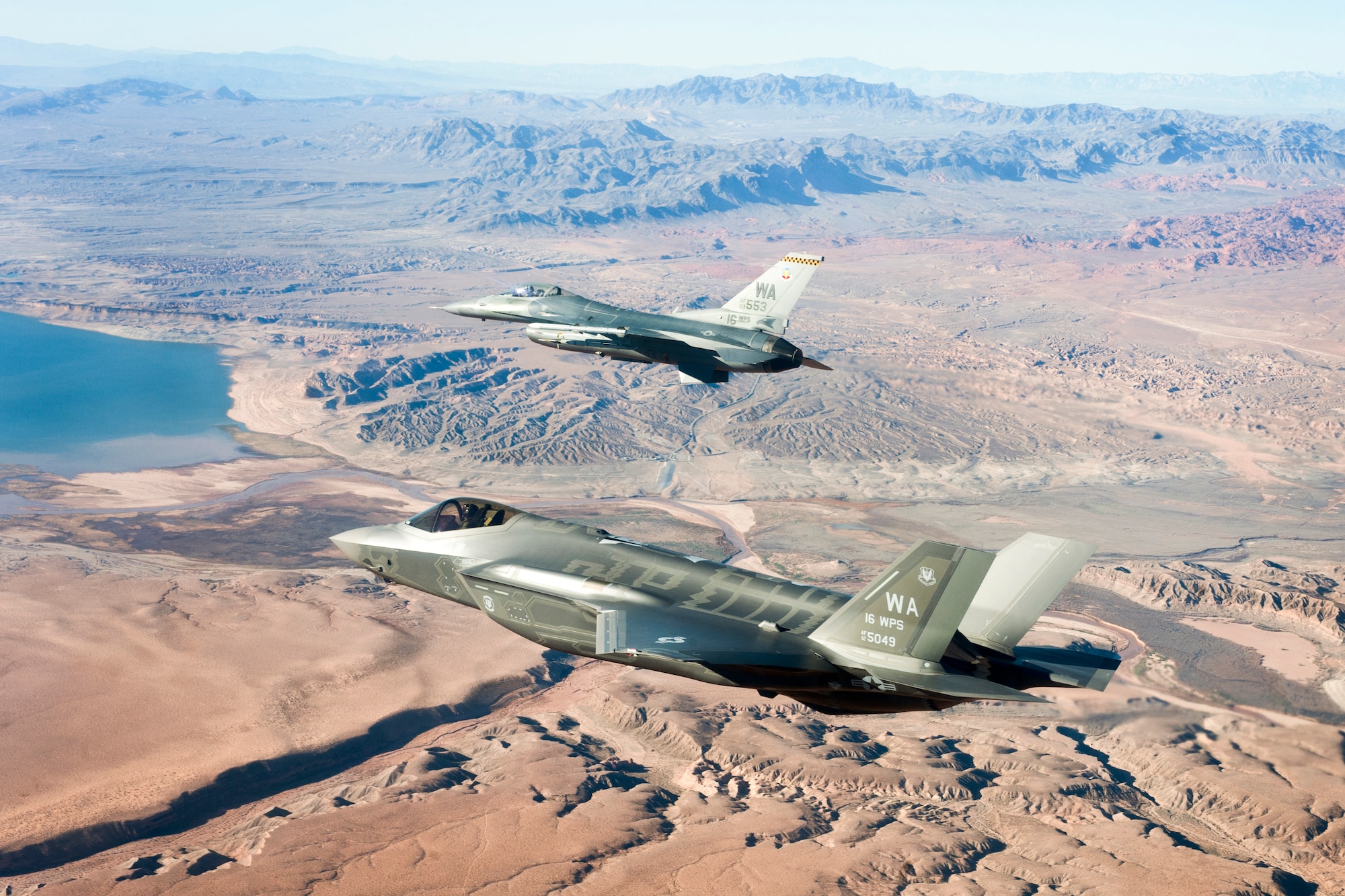 An F-35A Lightning II and F-16 Fighting Falcon assigned to the16th Weapons Squadron, U.S. Air Force Weapons School approach the Lake Mead National Recreation Area in route to Nellis Air Force Base, Nev., from the Lockheed Martin plant, Fort Worth, Texas, Jan. 15, 2015. The Weapons School's first F-35 will be used to drive tactics development and to create the curriculum for the first F-35 weapons instructor course.  (U.S. Air Force photo by Senior Airman Cody Griffith)