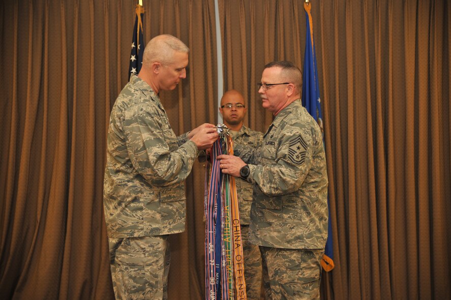(Center) Tech. Sgt. Marcus Young, executive assistant to the 319th Air Base Wing command chief, holds the wing’s guidon as Col. Pal Bauman, 319th ABW commander, pins on an Outstanding Unit Award streamer, while Command Chief Master Sgt. David Duncan stands by opposite of the colonel during a wing quarterly awards ceremony held Jan. 15, 2015, at the Northern Lights Club on Grand Forks Air Force Base, N.D. The Air Force Outstanding Unit Award is awarded by the secretary of the Air Force to numbered units that have distinguished themselves by exceptionally meritorious service or outstanding achievement that clearly sets the unit above and apart from similar units. (U.S. Air Force photo/Senior Airman Xavier Navarro)