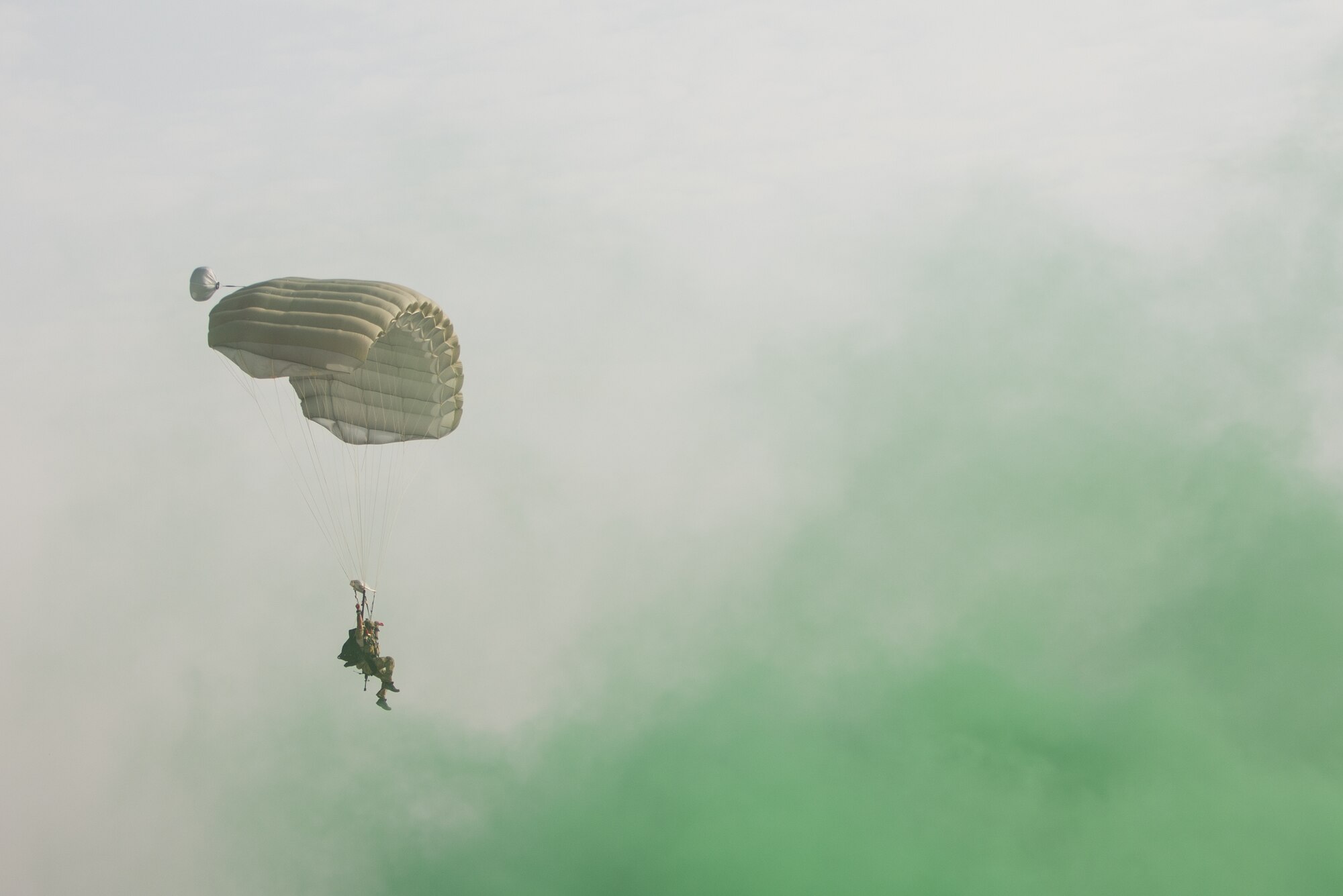 A U.S. Air Force Pararescueman with a Guardian Angel team assigned to the 83rd Expeditionary Rescue Squadron parachutes to the ground during High Altitude Low Opening jump training at Bagram Airfield, Afghanistan, July 18, 2015. To mark the drop zone, green smoke was used to guide the Pararescuemen to their landing point. The 83rd ERQS mission is to rescue, recover, and return American or allied forces in times of danger or extreme duress. (U.S. Air Force photo by Tech. Sgt. Joseph Swafford/Released)